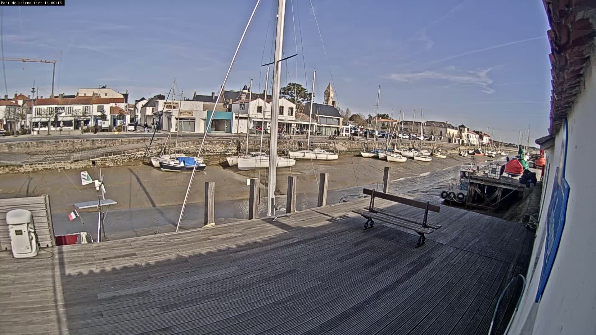 A sunny day shows sailboats docked in a harbor with a low tide exposing the muddy bottom, and a wooden dock with a bench in the foreground.