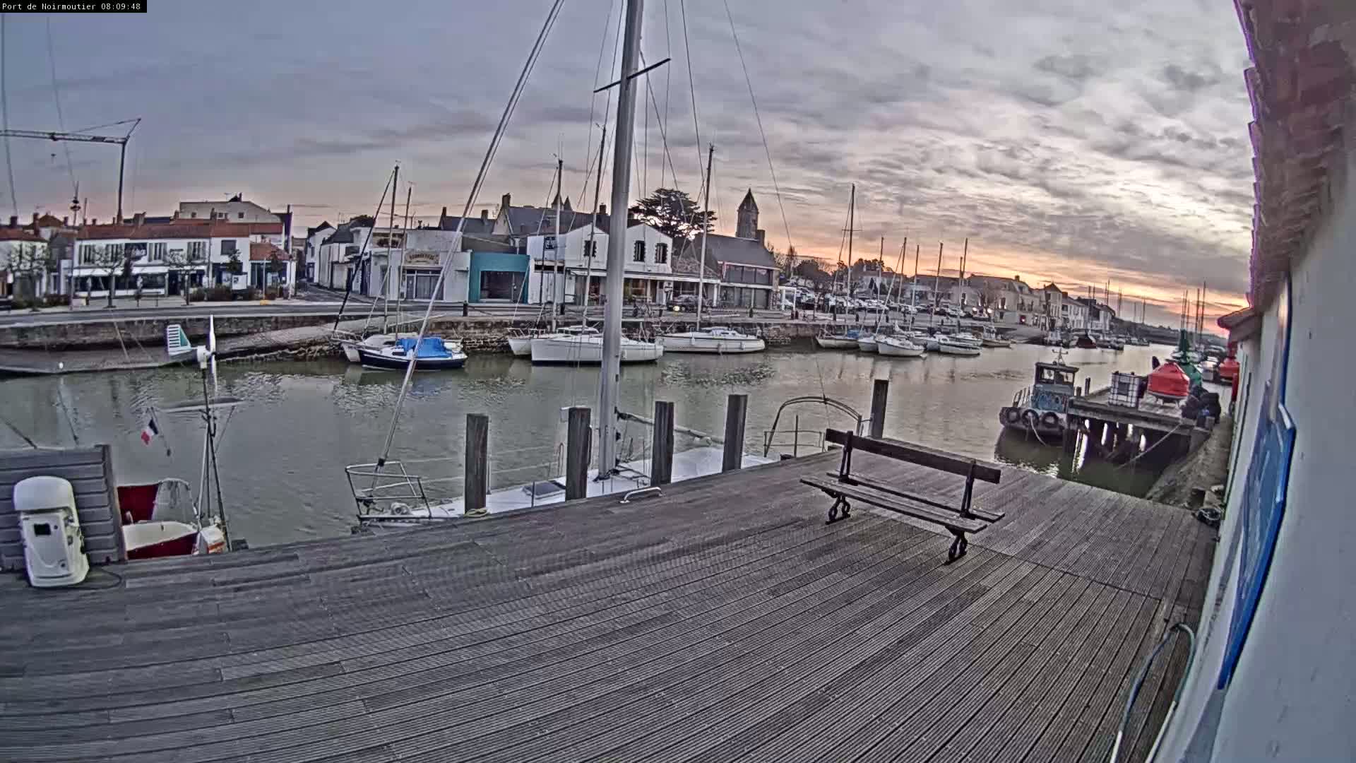 A harbor filled with sailboats at sunset under a cloudy sky, viewed from a wooden dock with a bench.