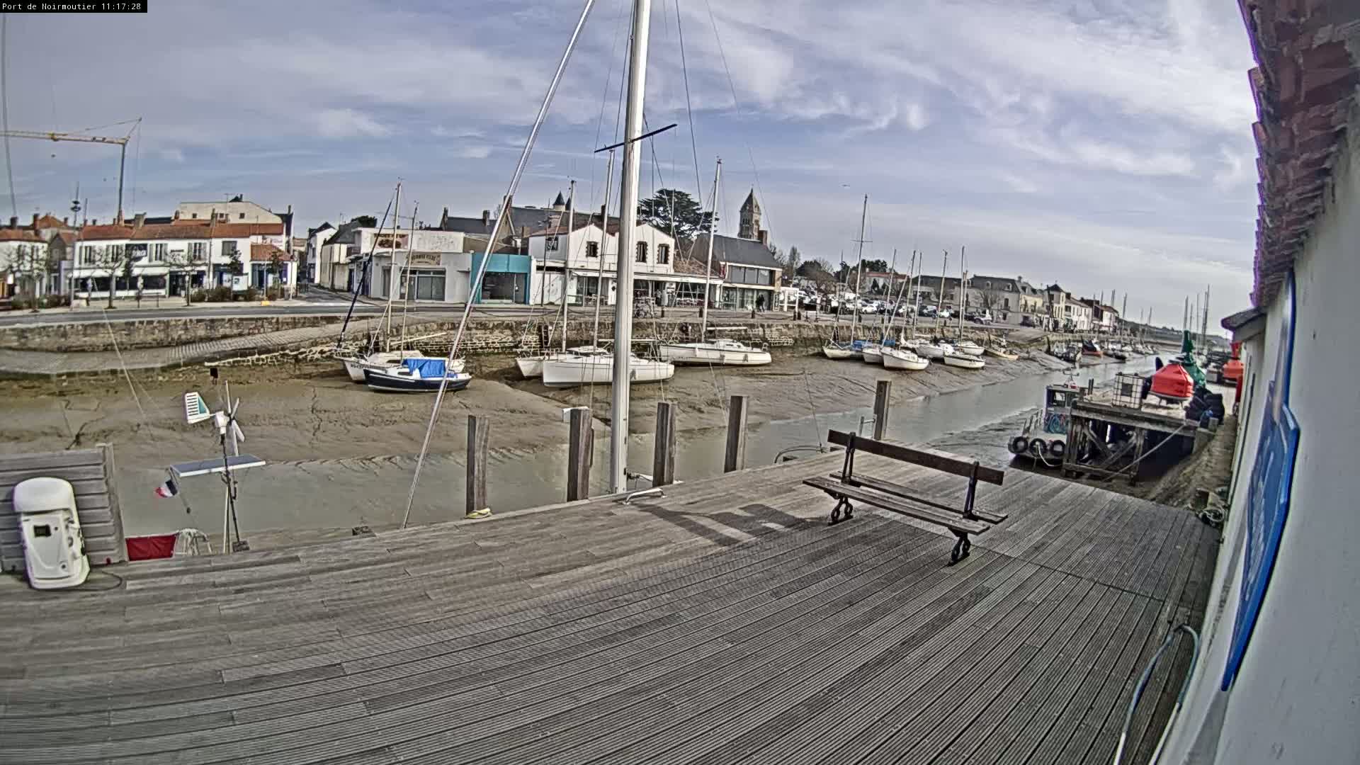 A mostly sunny day reveals sailboats docked in a harbor at low tide, with a wooden dock in the foreground and a small town visible in the background.