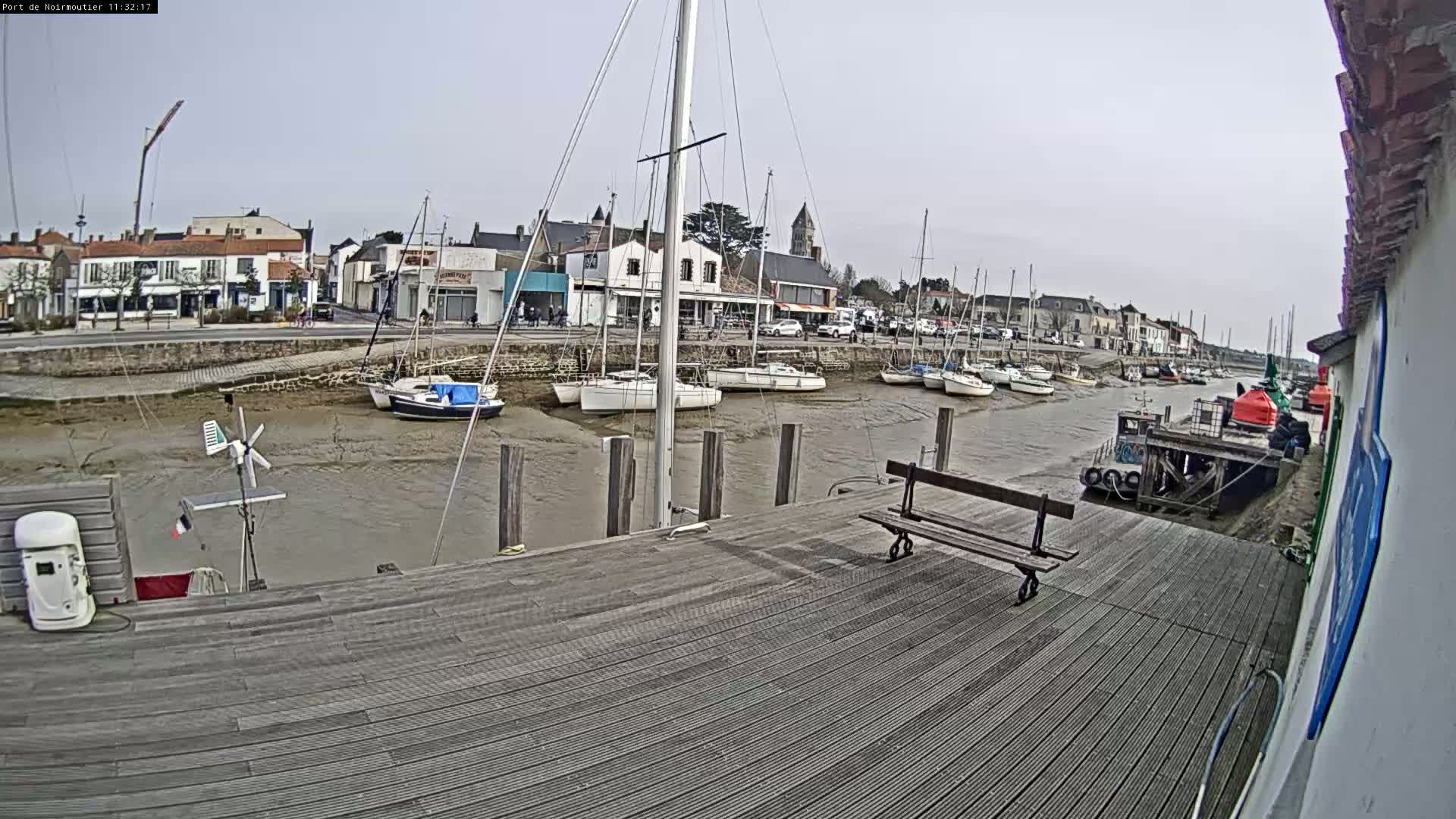 A wooden dock overlooks a calm harbor with several sailboats at low tide under an overcast sky.