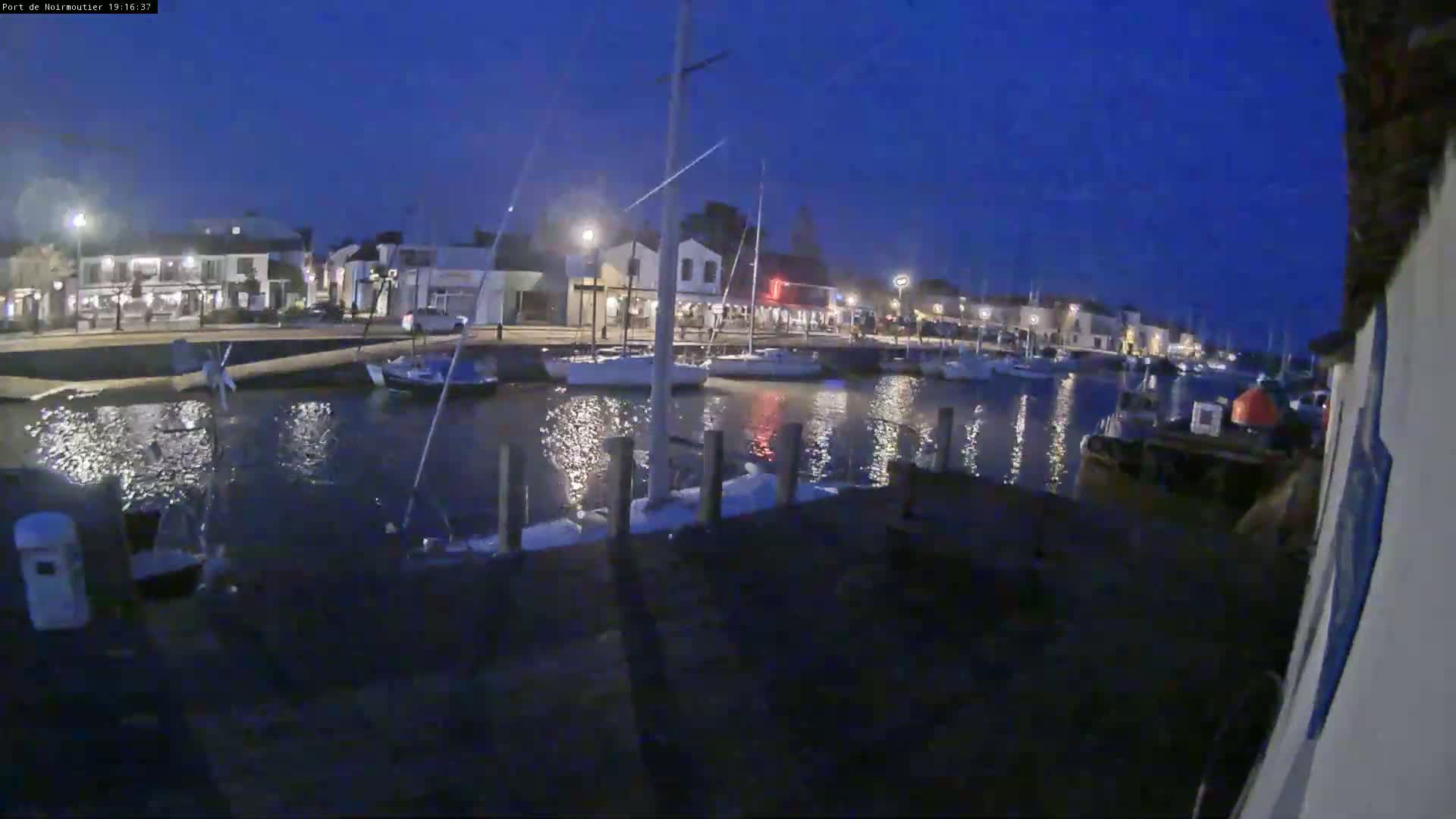 A calm harbor at night, with sailboats moored along a waterfront lined with buildings, under a twilight sky.