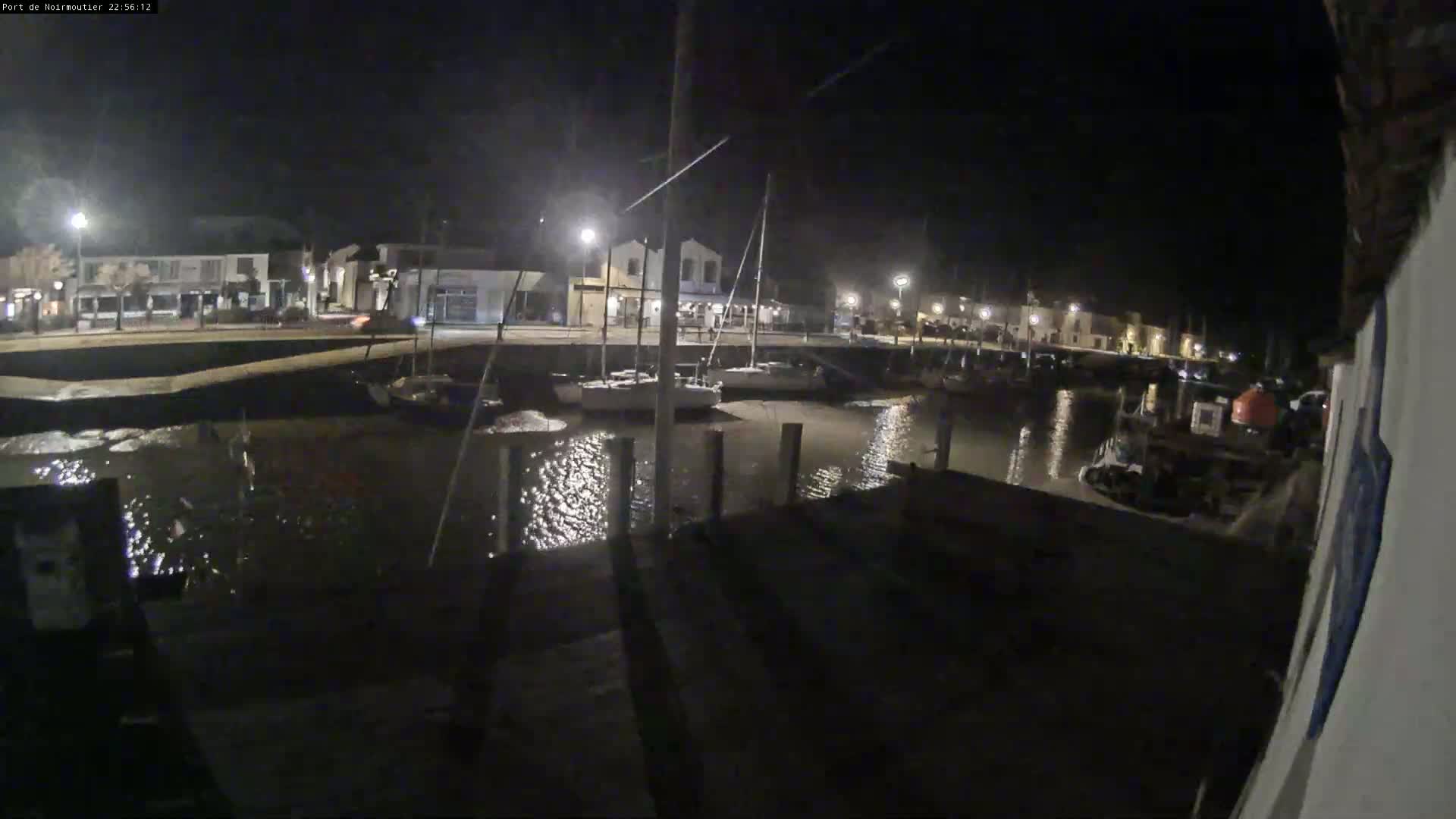 At night, several boats are moored in a calm harbor next to a waterfront village illuminated by streetlights.