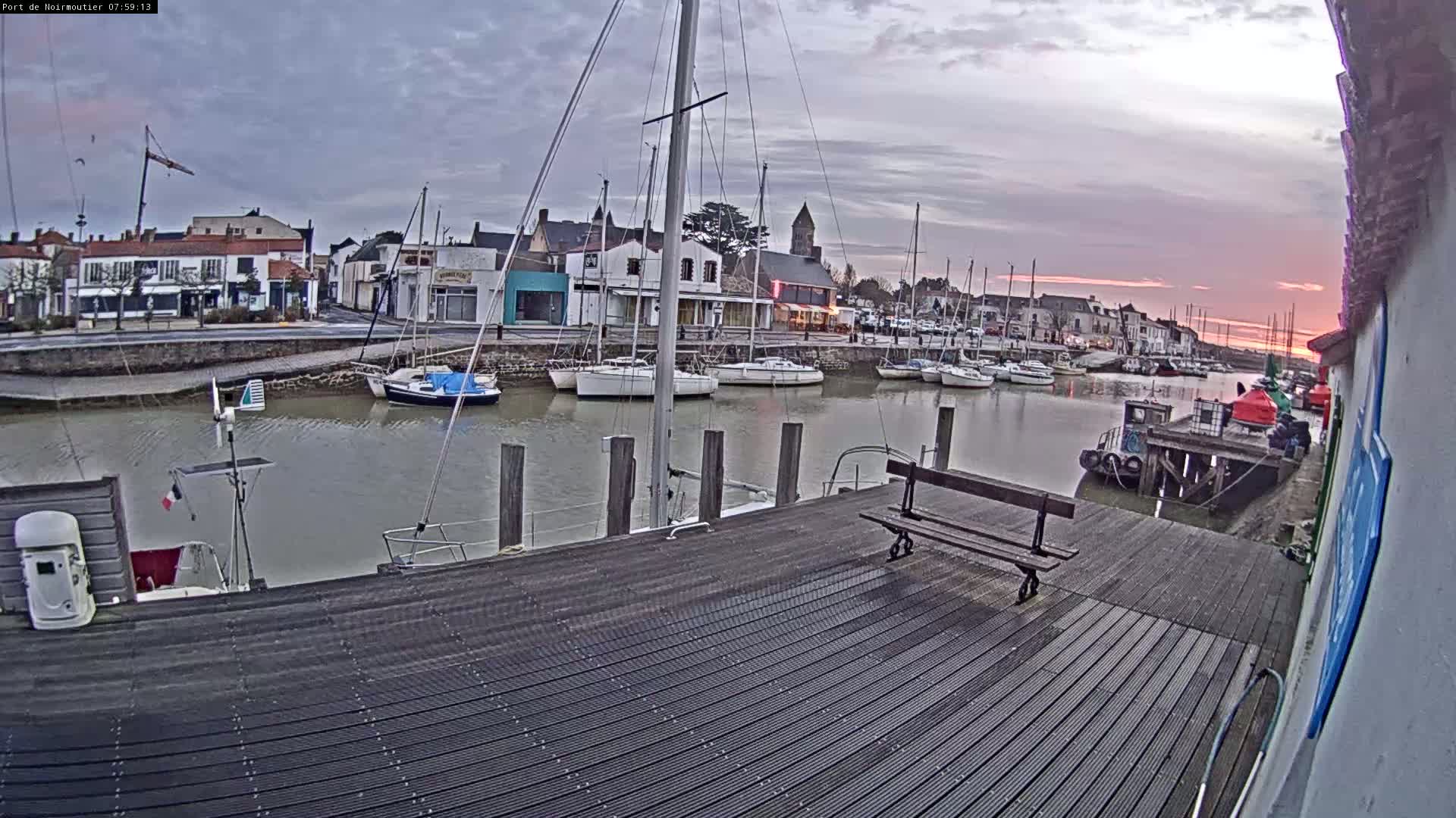 A harbor filled with sailboats at dawn under a cloudy sky, viewed from a wooden dock with a bench.