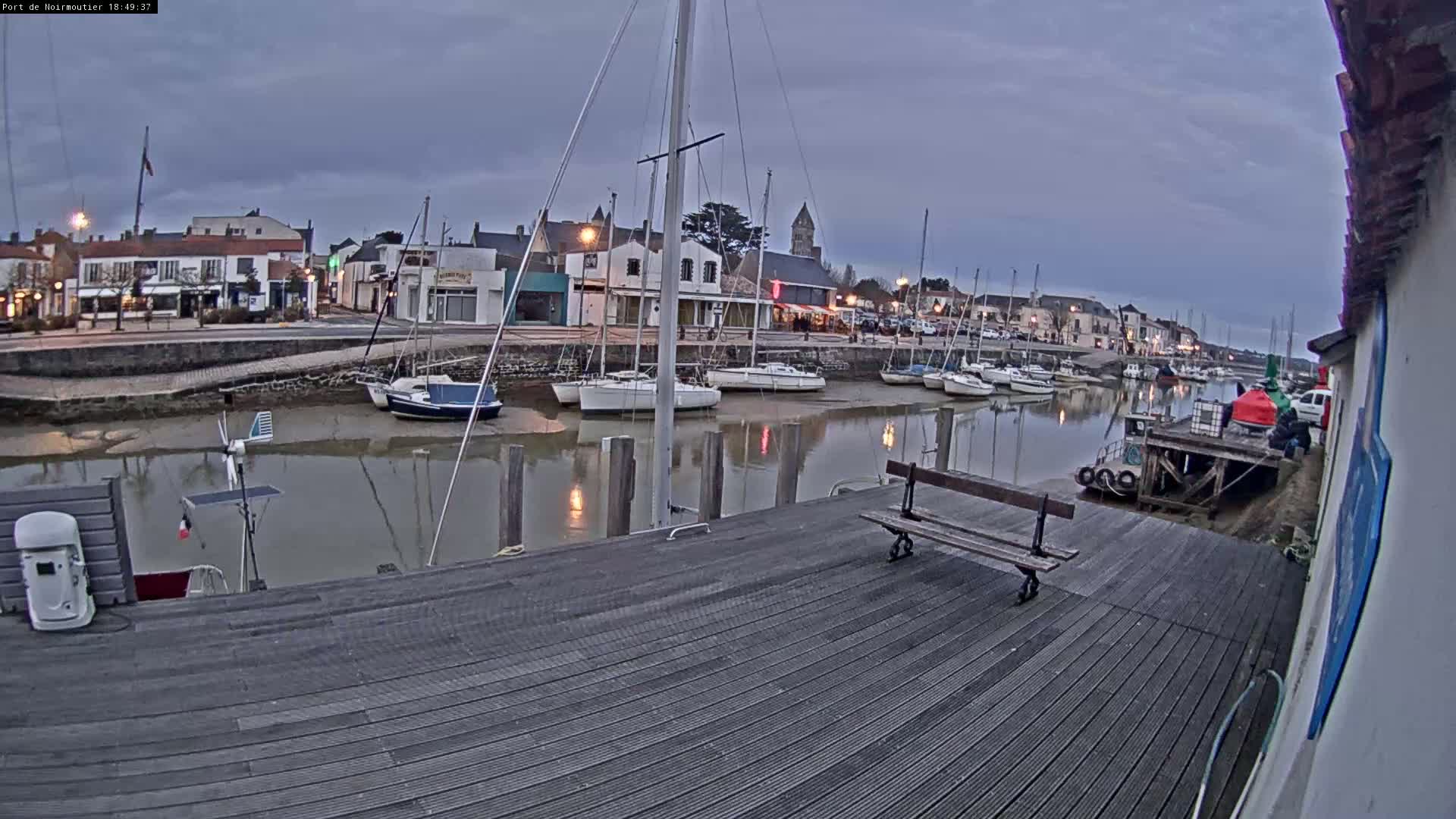 A wooden dock overlooks a calm harbor filled with sailboats at low tide under an overcast sky.