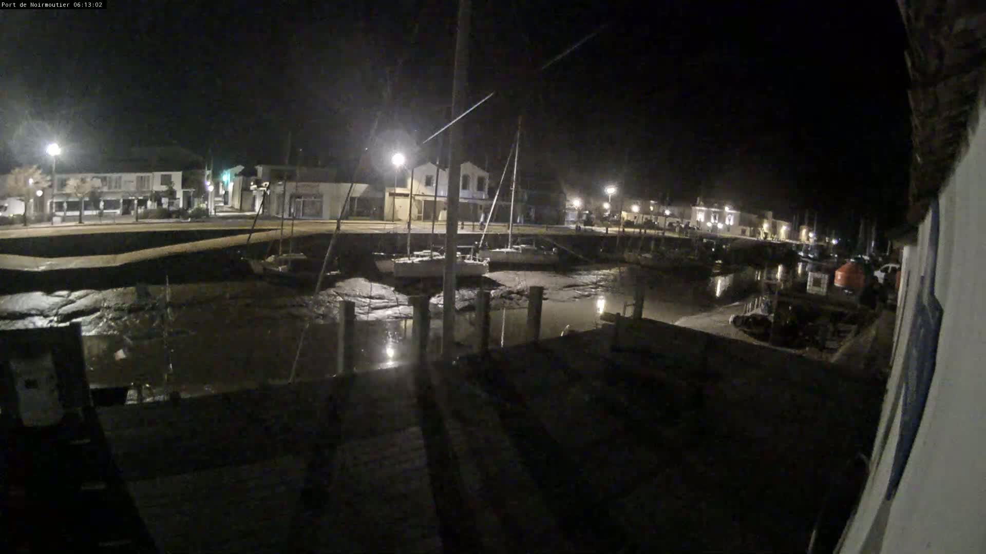 At night, several sailboats are docked in a harbor with low water levels, illuminated by streetlights along a waterfront building.