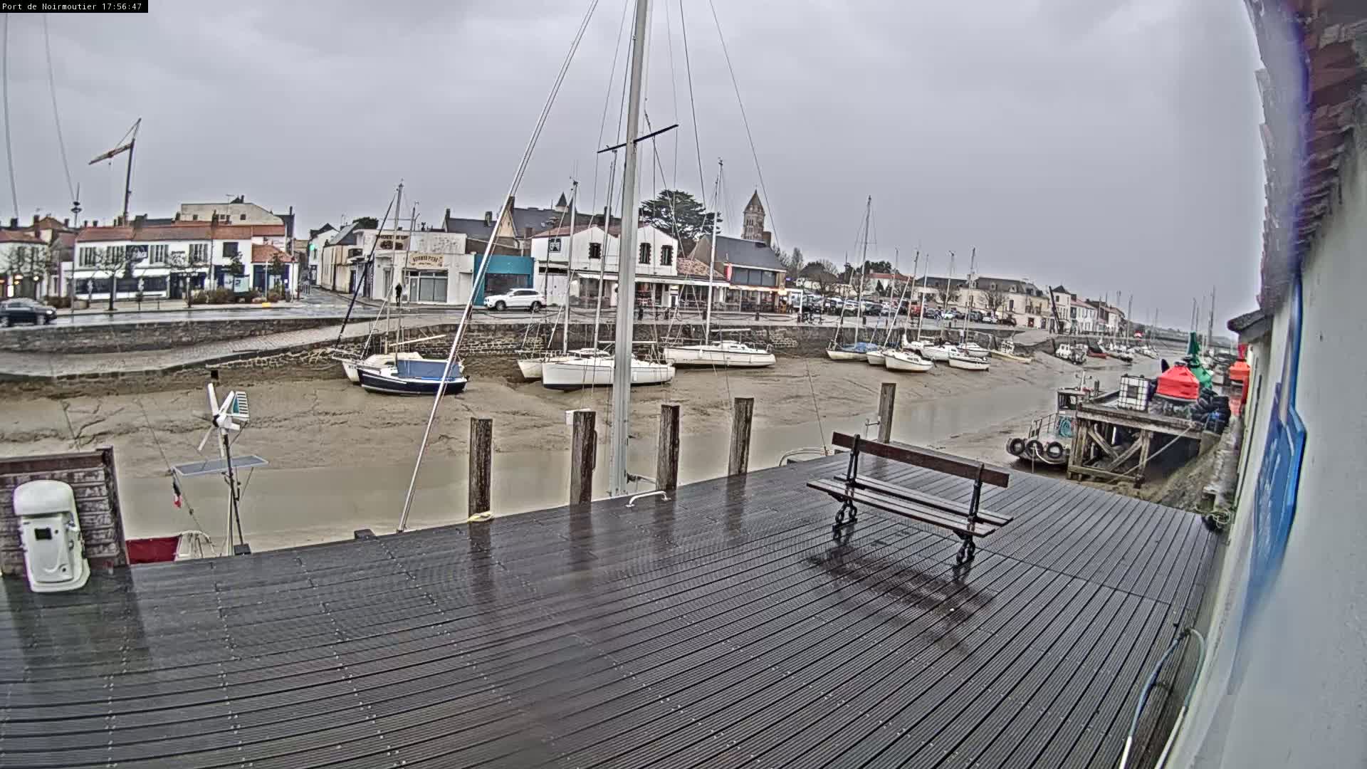 A wet wooden dock overlooks a harbor with several sailboats at low tide under an overcast sky.