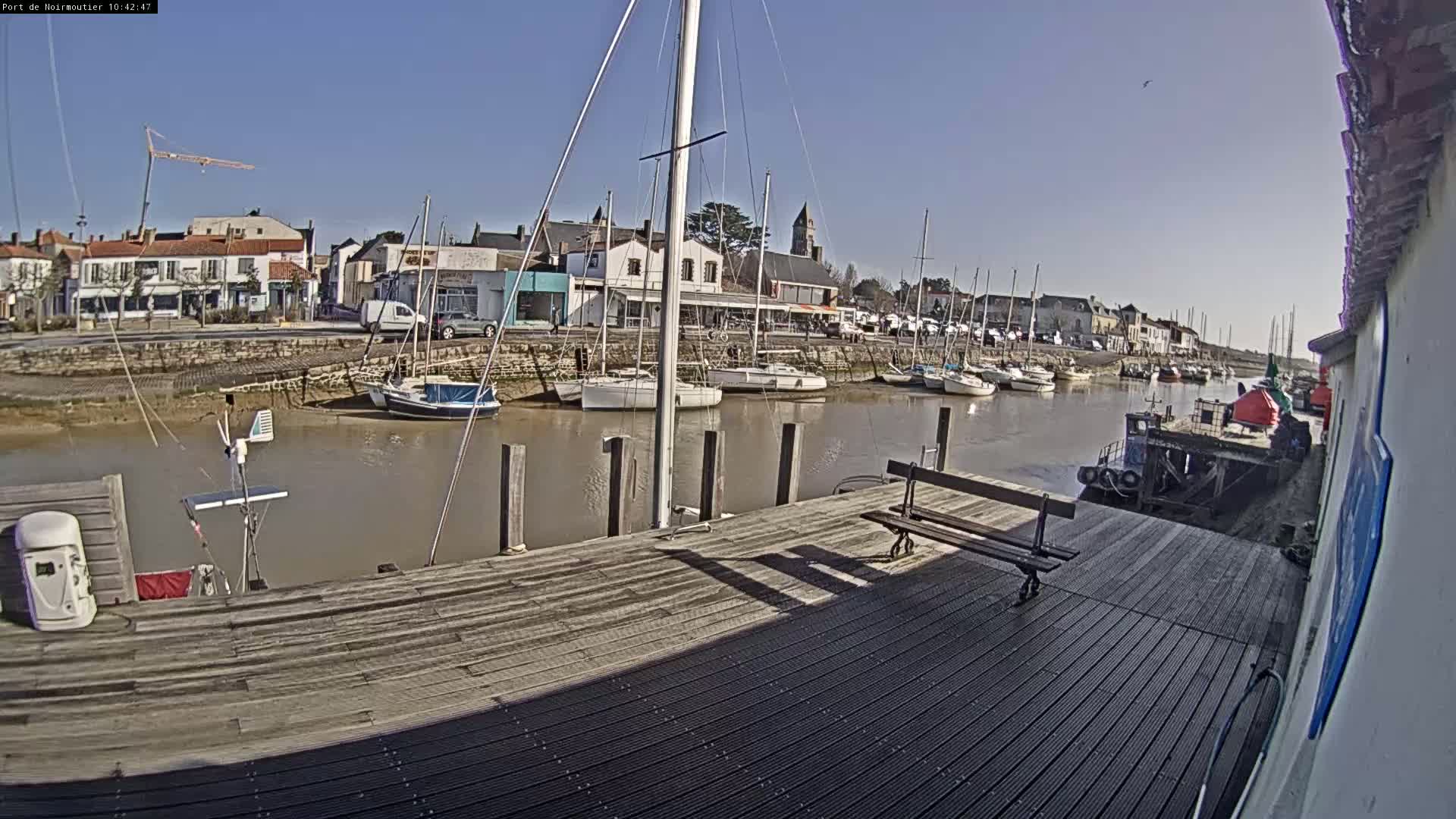 A sunny day at a harbor shows several sailboats moored along a calm waterway, with a wooden dock in the foreground and a small town visible beyond the water.