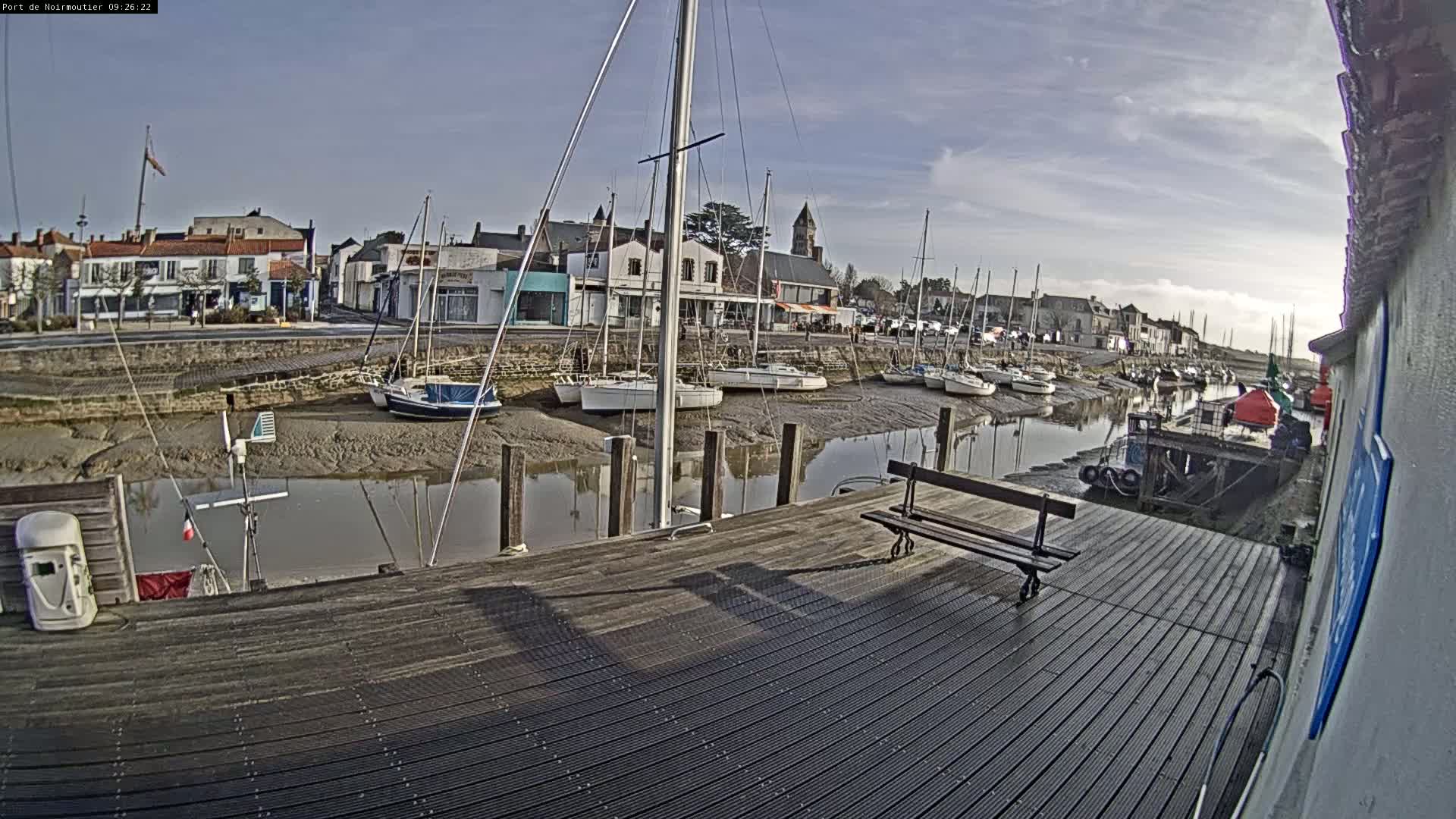 A mostly sunny day reveals a small harbor with sailboats docked along a muddy shore, viewed from a wooden deck with a bench.