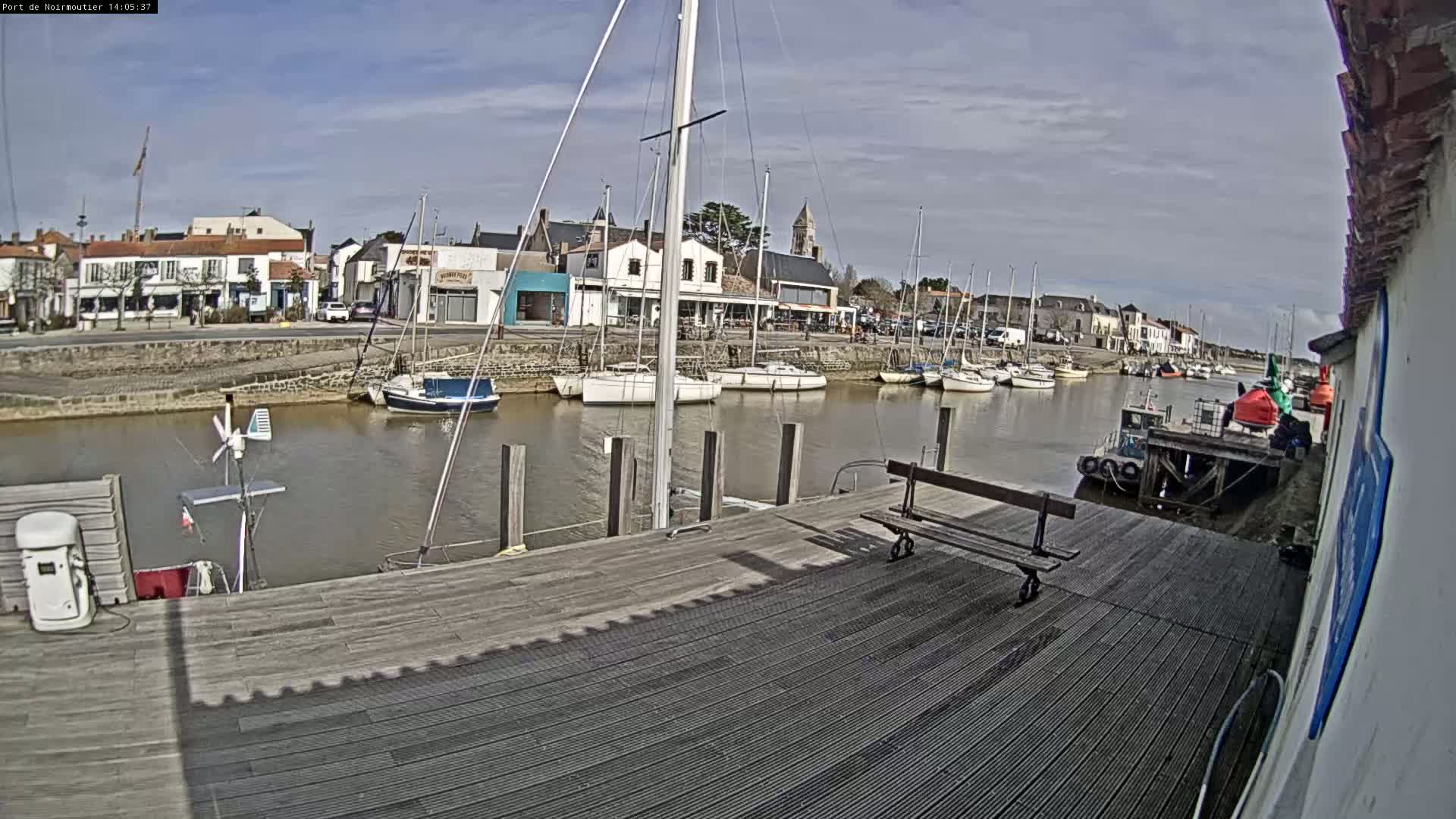 A mostly sunny day at a harbor shows several sailboats moored along a dock, with a town visible in the background.