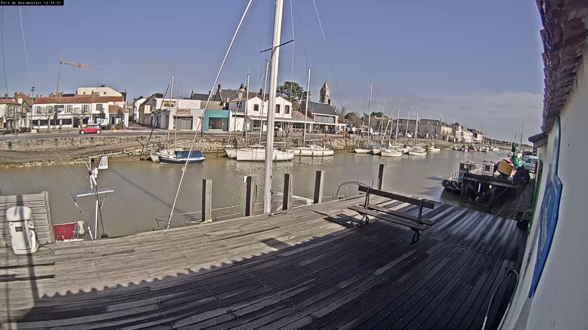 A wooden dock extends over calm water, past moored sailboats and a small town under a clear, sunny sky.
