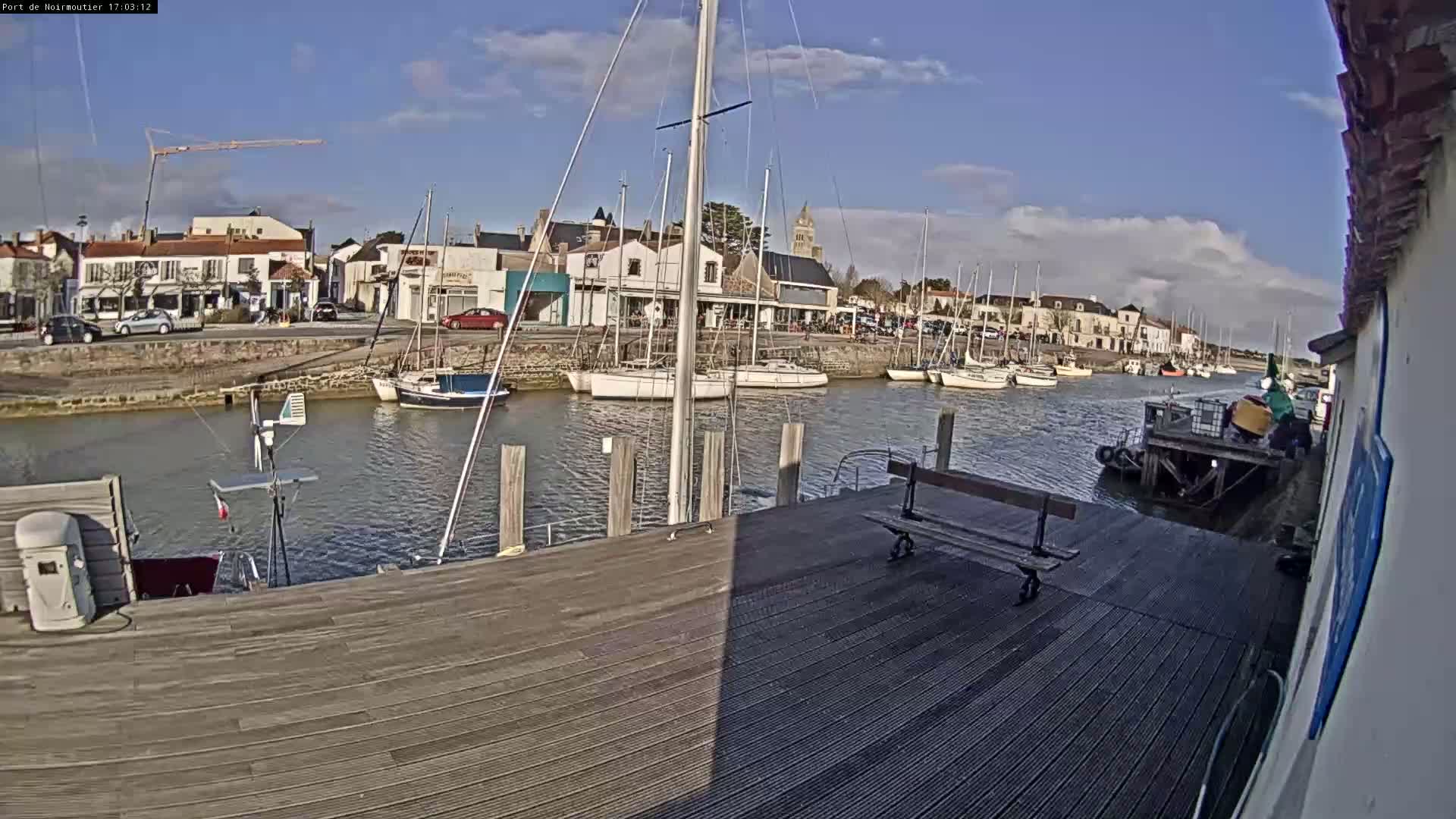 A mostly sunny day at a harbor shows sailboats moored along a quay with a wooden dock in the foreground and a village in the background.