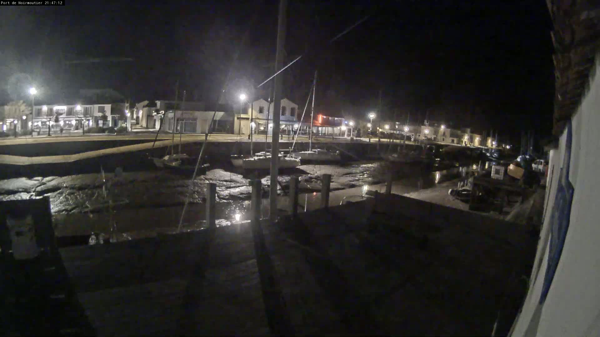 At night, a small harbor with boats on exposed mudflats is illuminated by streetlights, bordering buildings along a waterfront.