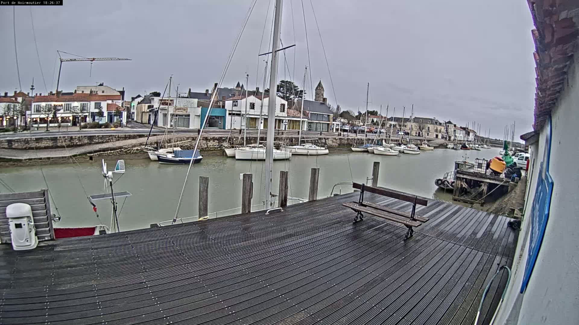 A harbor with several sailboats docked, a wooden dock with a bench, and overcast skies.