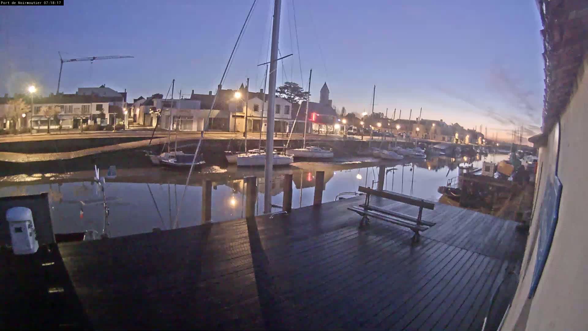 A calm harbor at dawn, with sailboats moored along a dock next to a town's waterfront buildings.