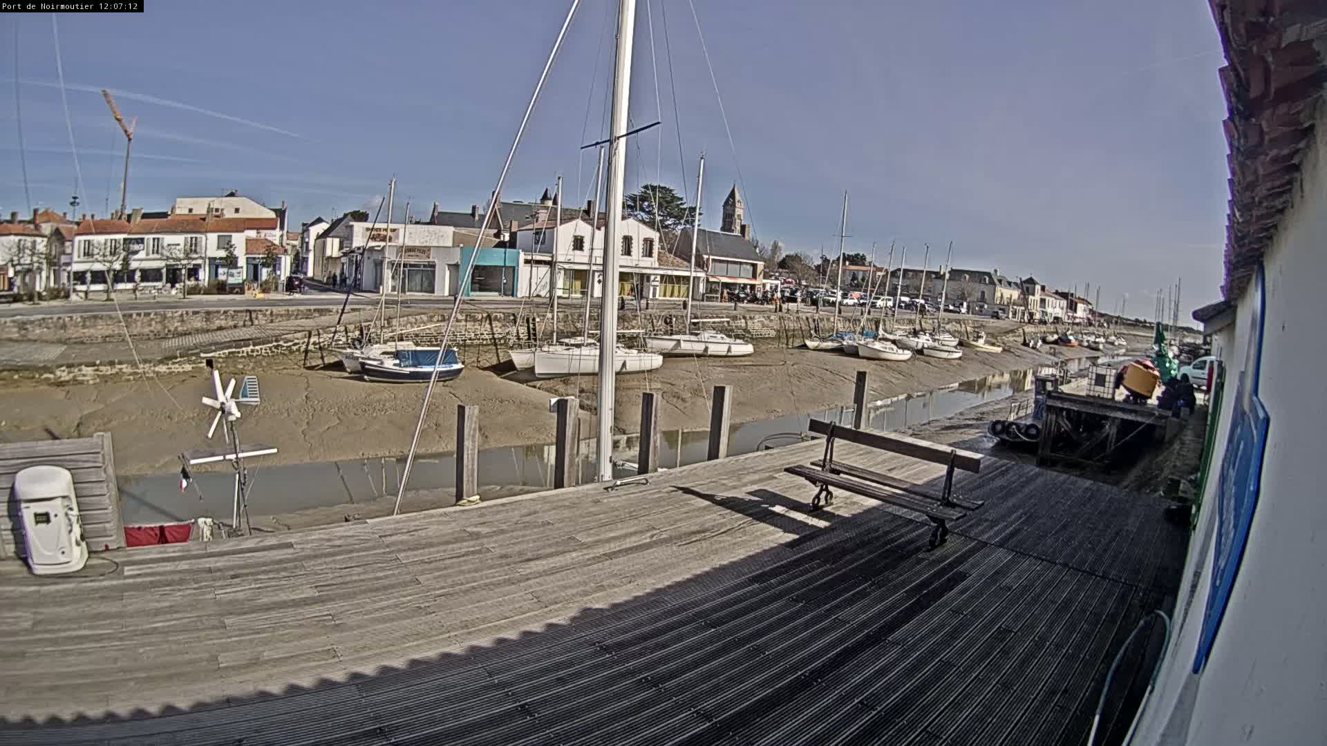 A wooden dock overlooks a calm waterway at low tide, where numerous sailboats are grounded on the muddy shore in front of a small town under a clear, sunny sky.