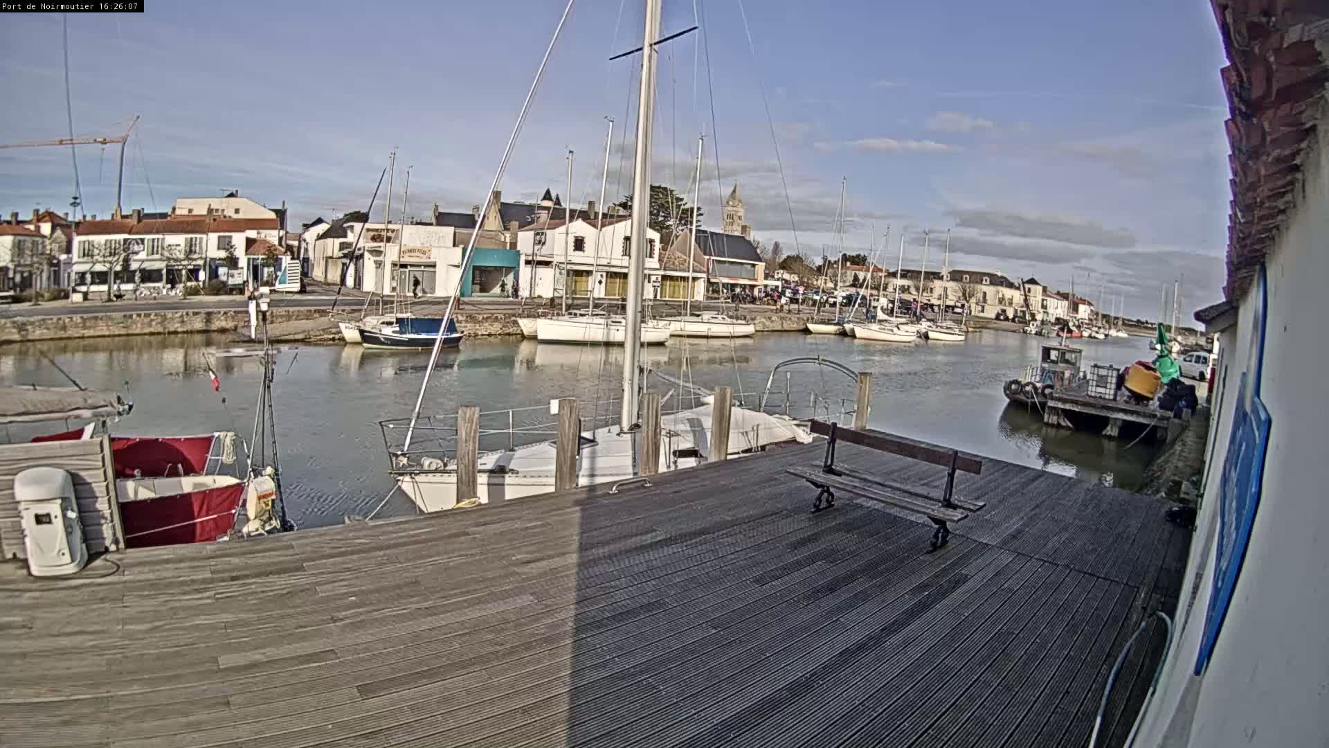 A sunny day at a harbor shows numerous sailboats docked along a waterfront lined with buildings, a wooden dock in the foreground, and a calm body of water.