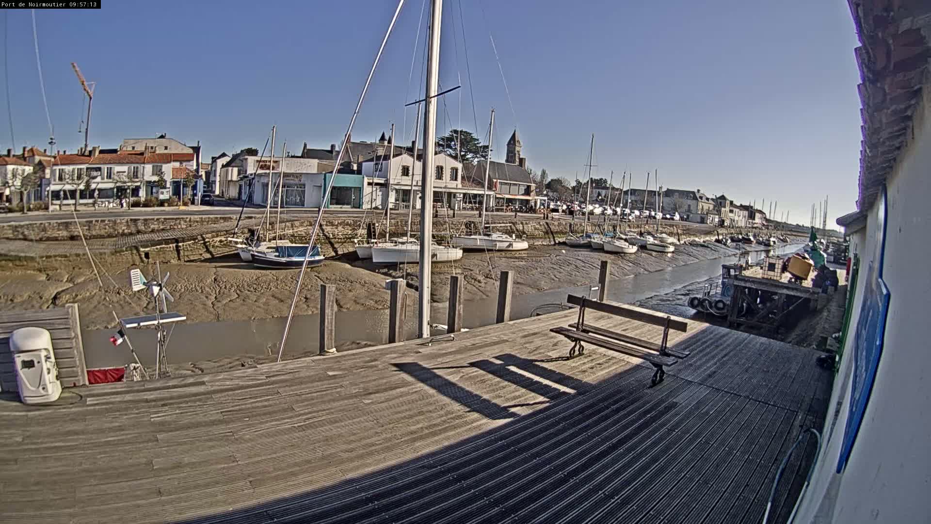 A wooden dock overlooks a narrow waterway at low tide, where several sailboats are grounded on the mudflats near a small waterfront town under a clear, sunny sky.