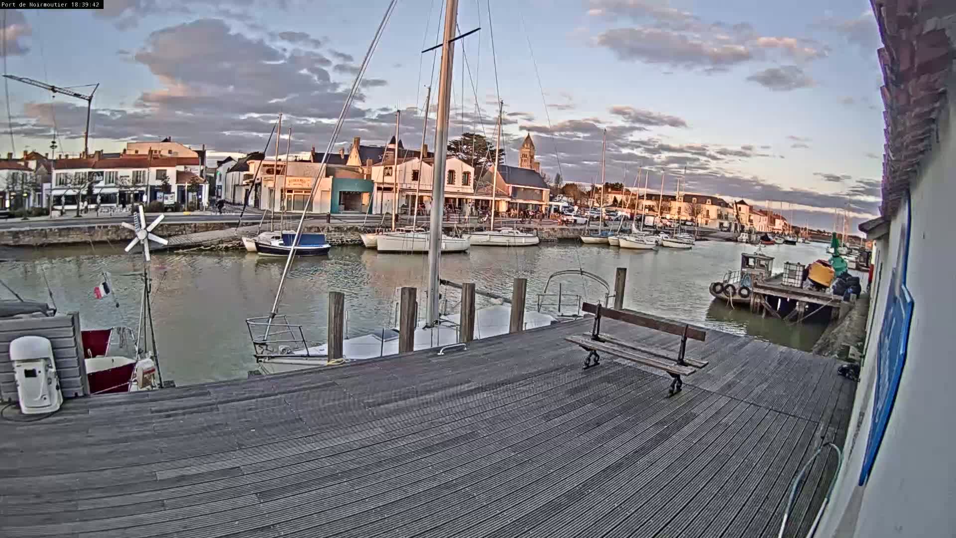A harbor filled with sailboats under a partly cloudy evening sky, viewed from a wooden dock.