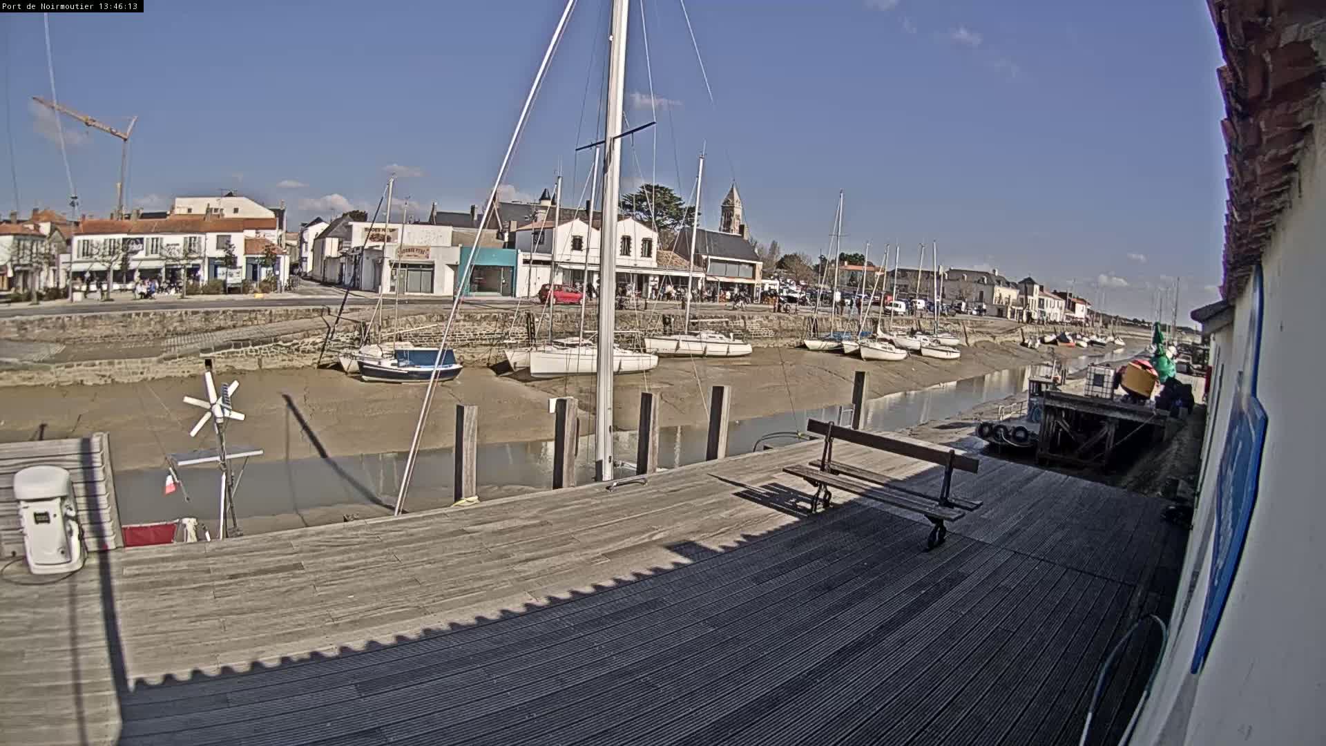 A sunny day at a harbor shows several sailboats docked at low tide, with a wooden dock in the foreground and a small town visible in the background.