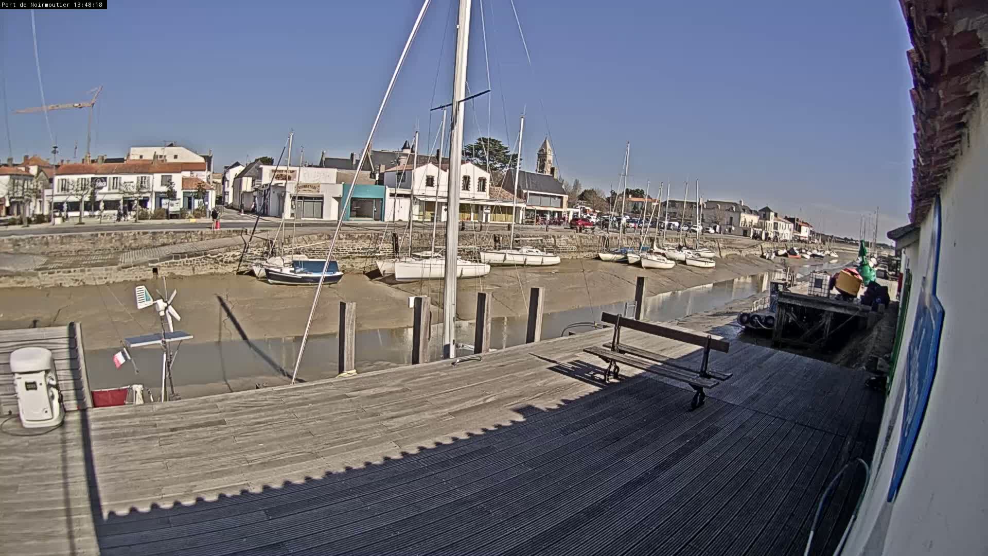 A sunny day shows sailboats docked in a shallow waterway next to a village with a wooden dock in the foreground.
