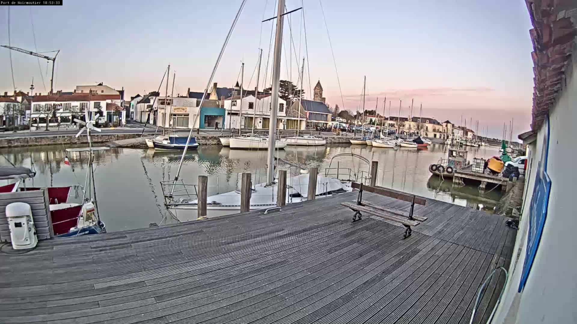 A calm harbor filled with sailboats at dusk, viewed from a wooden dock with a bench.