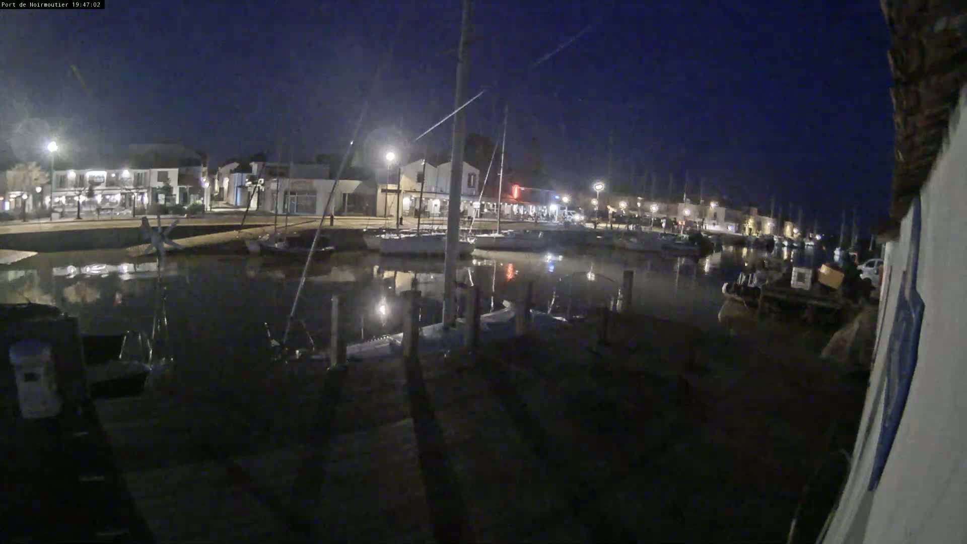 At night, a calm harbor filled with boats is illuminated by lights from buildings along the waterfront.