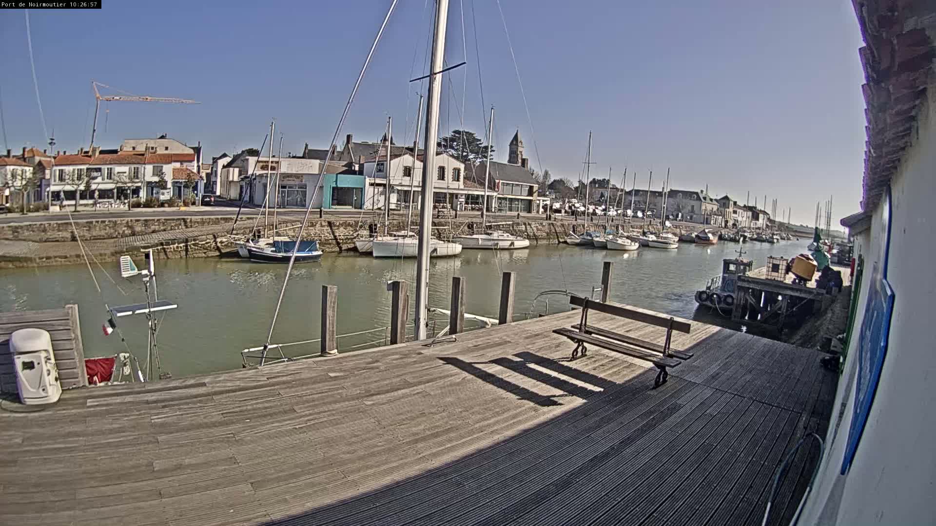 A sunny day at a harbor shows sailboats docked along a calm waterway, with a wooden dock extending towards the viewer and a small town visible in the background.