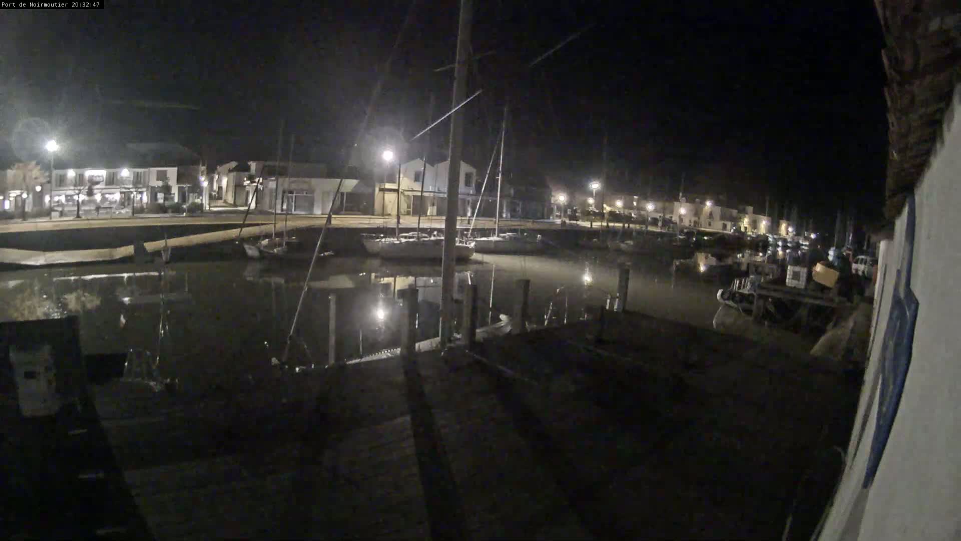 At night, a harbor is shown with boats moored alongside a dock, and buildings with lights are visible in the background.