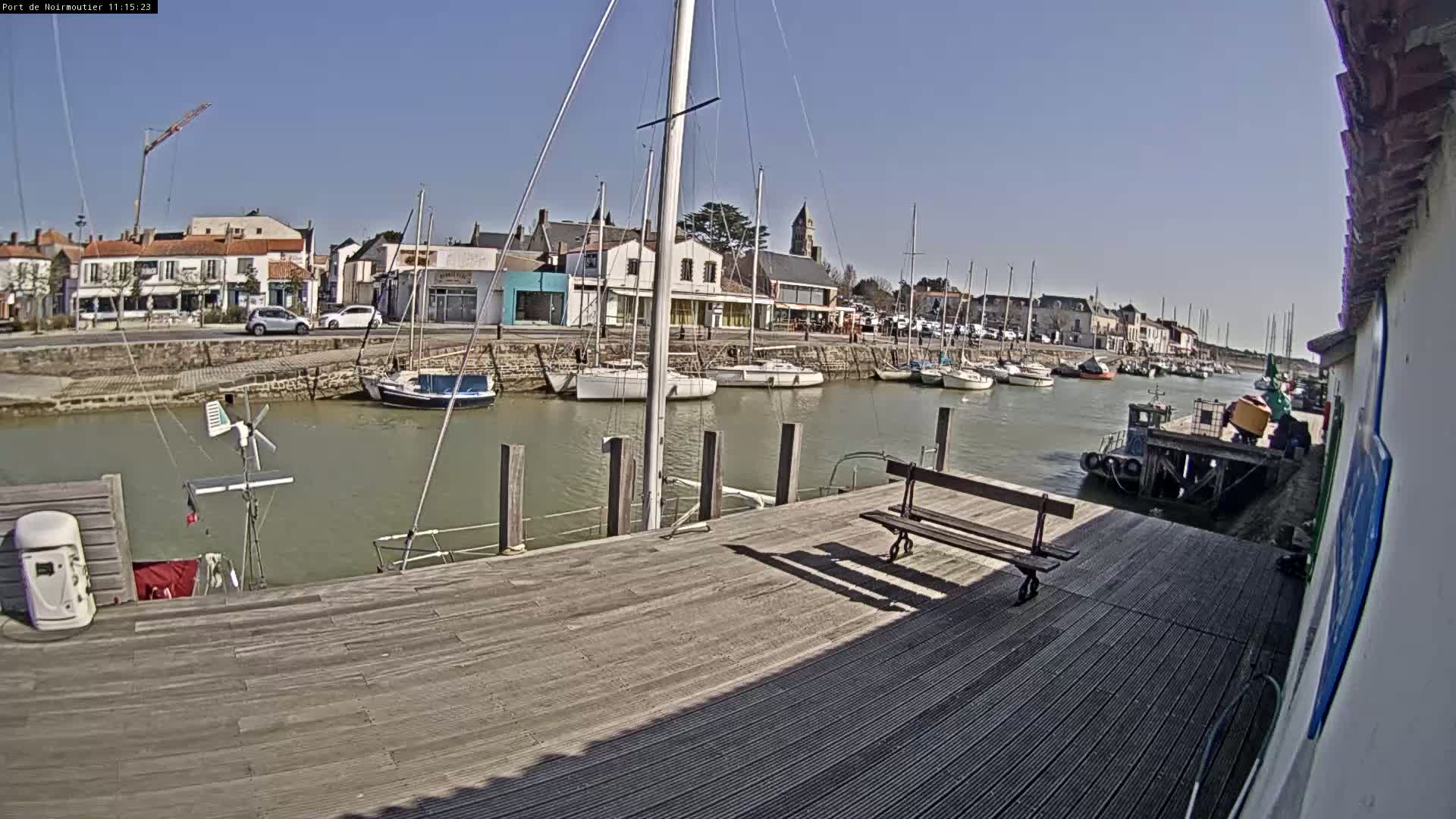 A sunny day at a harbor shows sailboats docked along a waterway next to a town with buildings and cars.