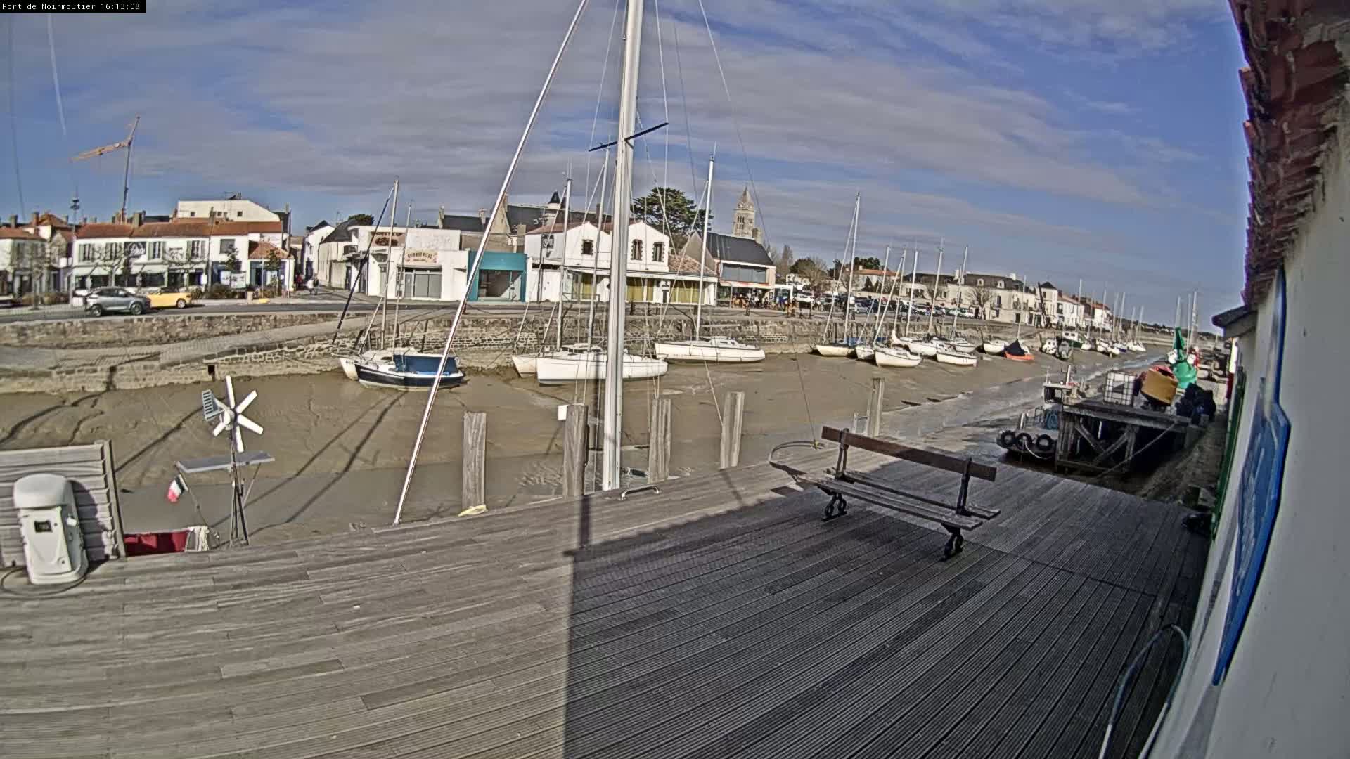 A mostly sunny day reveals a harbor with several sailboats docked at low tide, beside a wooden dock and a small town in the background.