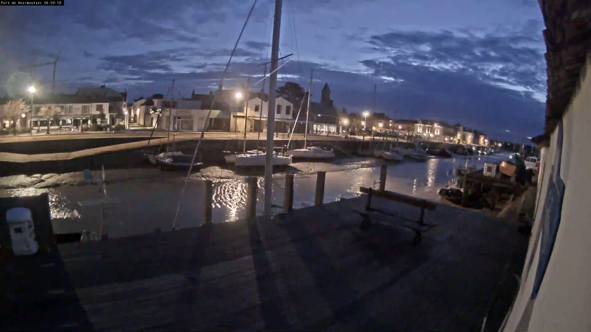A nighttime view of a harbor with sailboats docked, a calm body of water, and a town illuminated by streetlights under a partly cloudy sky.