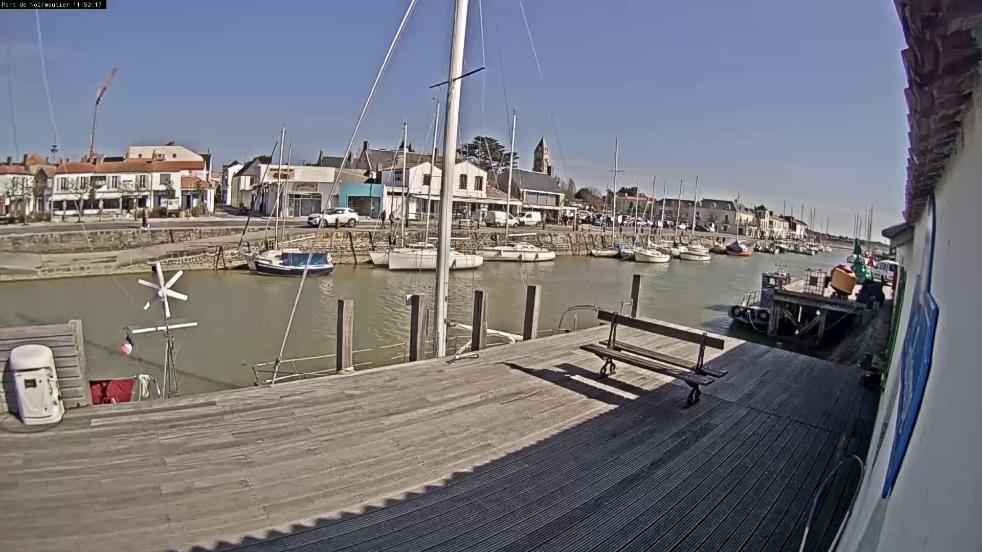 A sunny day at a harbor shows a wooden dock with several sailboats moored along a calm waterway, in front of a small town.