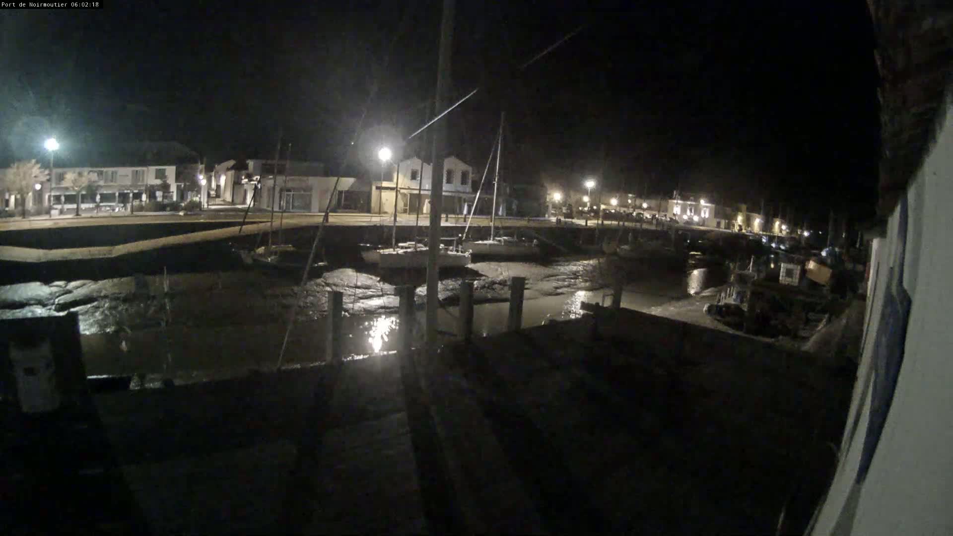 A nighttime view of a harbor with several boats docked, low tide exposing the muddy seabed, and buildings lit by streetlights in the background.