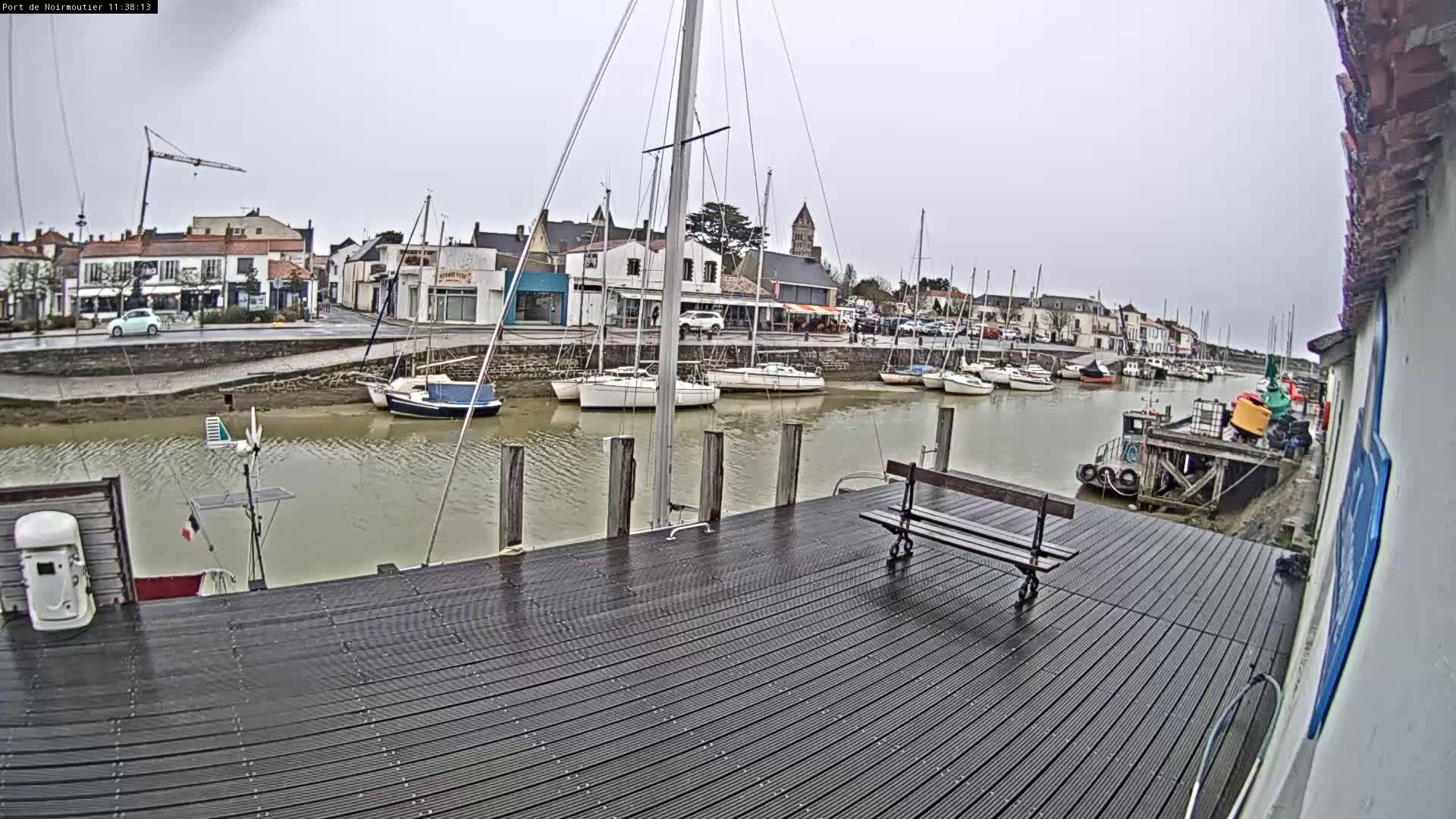 A wet wooden dock overlooks a calm harbor filled with sailboats moored alongside a waterfront town under an overcast sky.