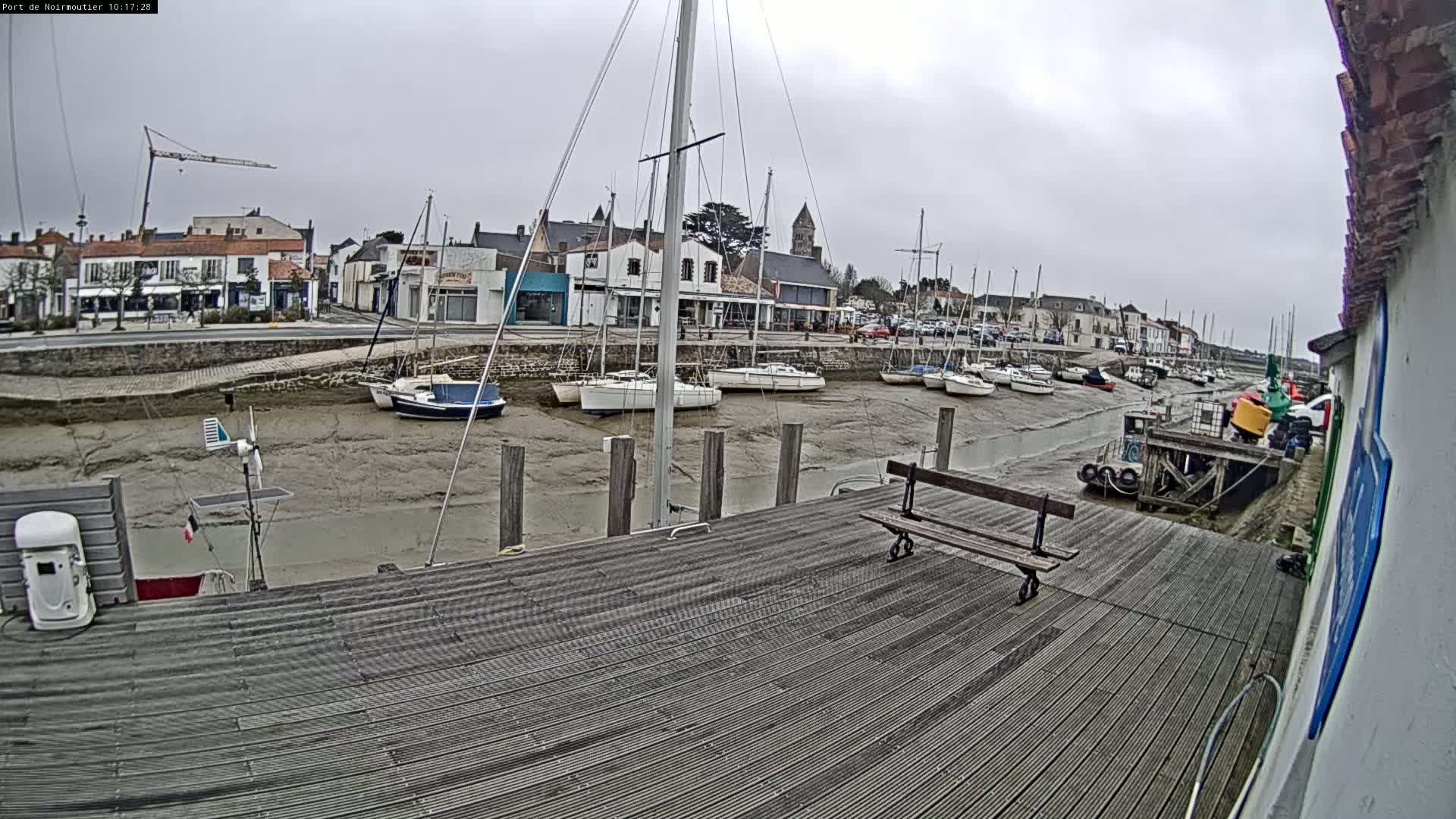 A wooden dock overlooks a harbor at low tide, where numerous sailboats are moored and a small town is visible in the background under an overcast sky.