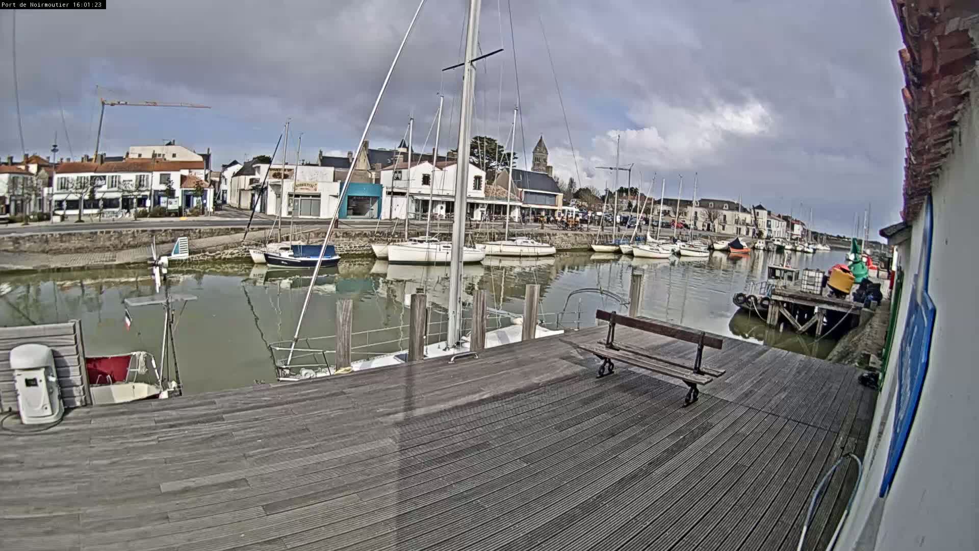A harbor with sailboats docked, under a cloudy sky, is viewed from a wooden dock.