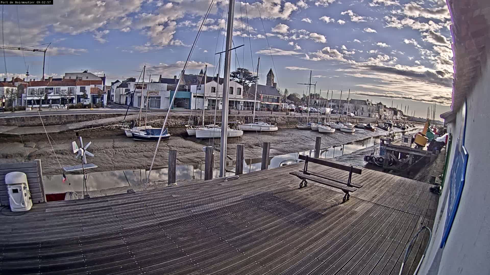 A wooden dock overlooks a harbor at low tide, where numerous sailboats are moored, under a partly cloudy sky.