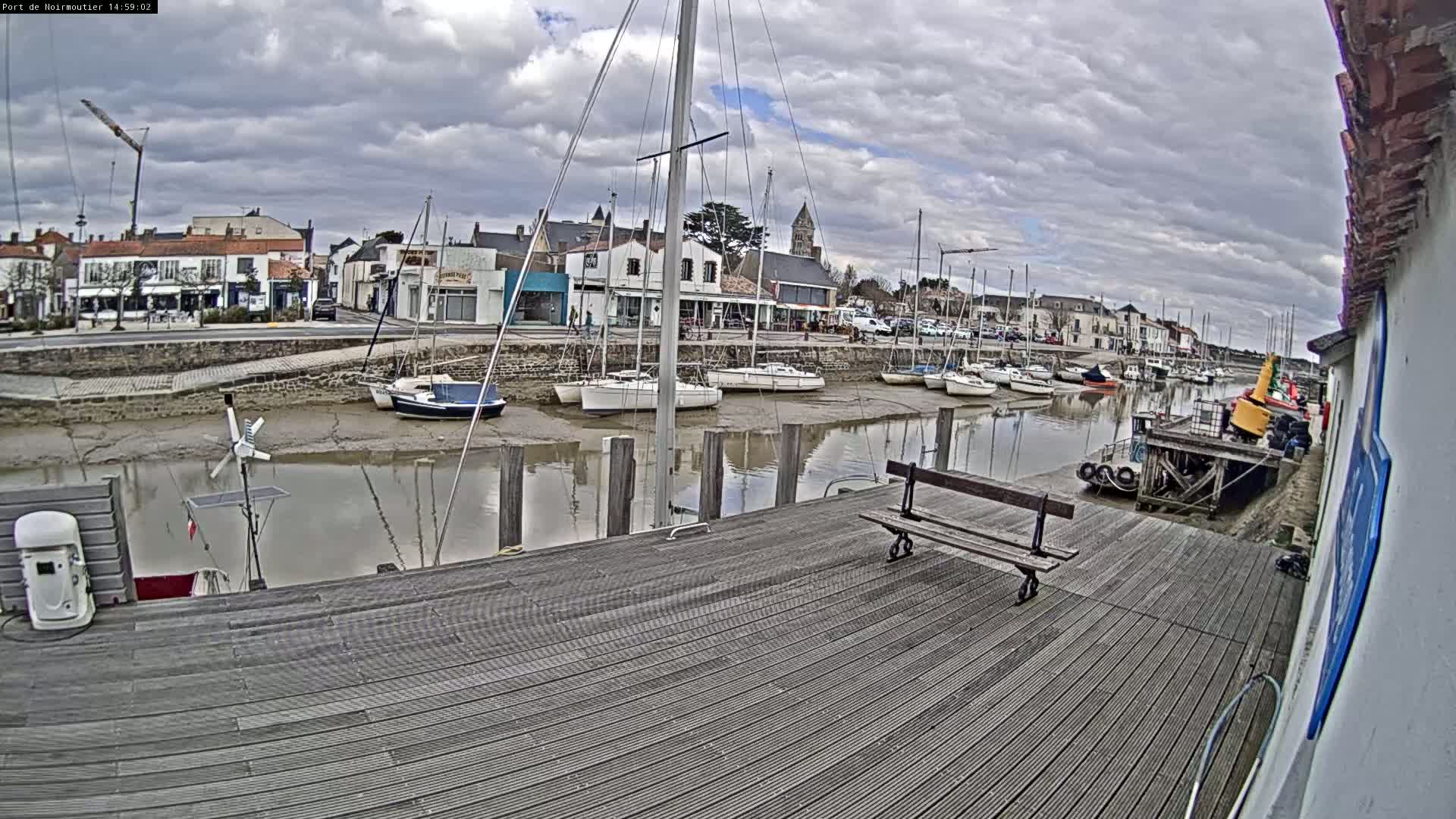 A wooden dock overlooks a calm waterway with several sailboats at low tide under a cloudy sky.