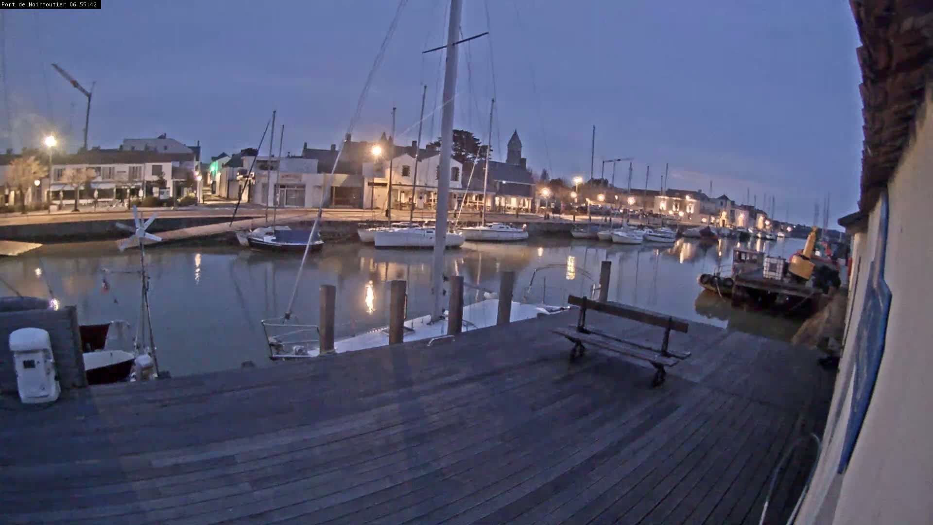 A wooden dock at twilight overlooks a calm harbor filled with sailboats and a small tugboat, under a clear, dark blue sky.
