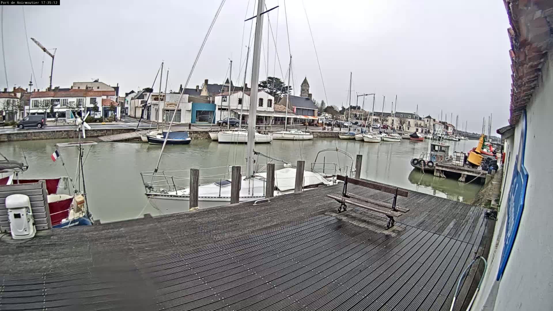 A wooden dock overlooks a calm harbor filled with sailboats and small motorboats under an overcast sky.