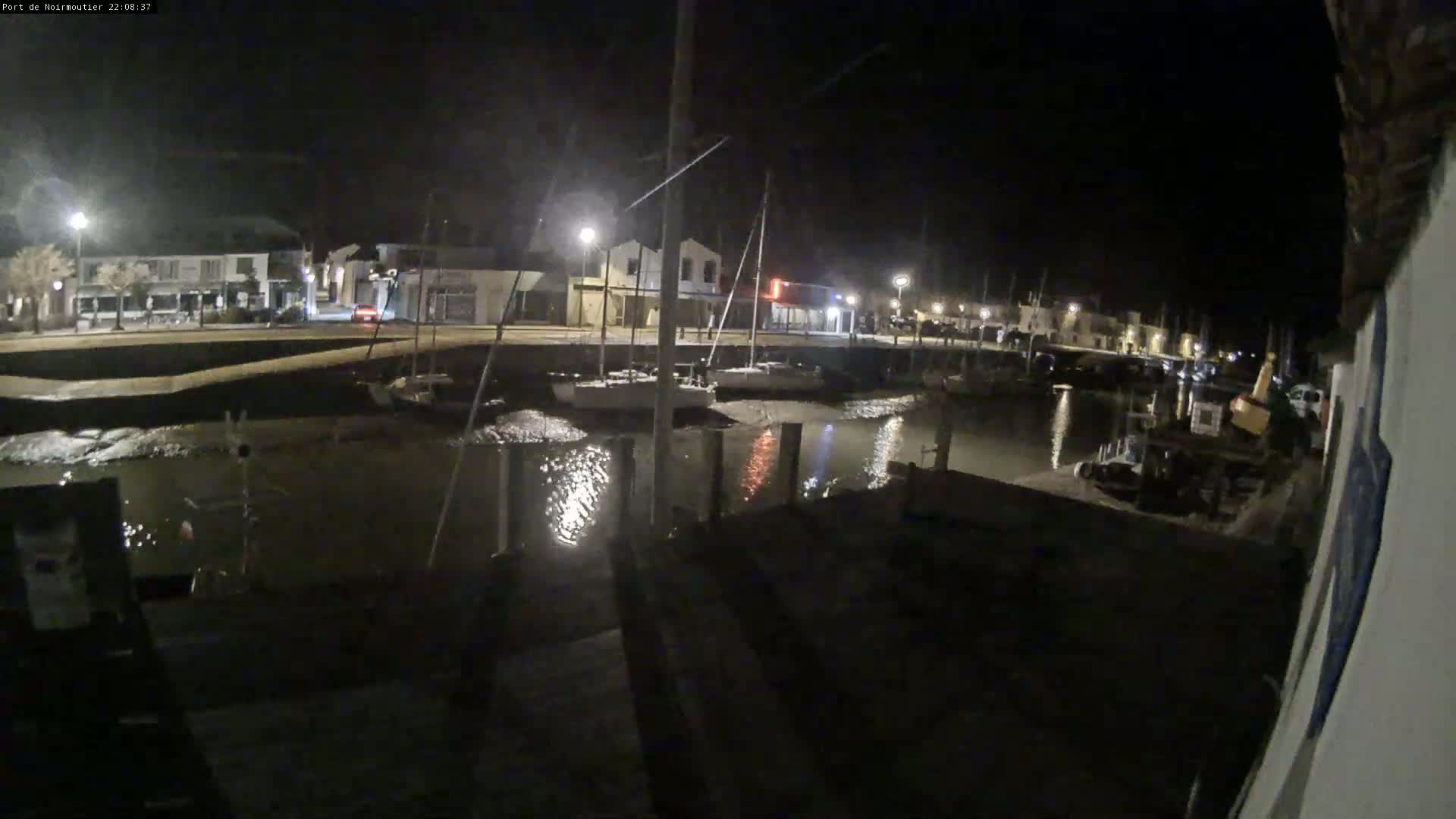 At night, a calm body of water reflects the lights of a small, waterfront town, where several sailboats are docked.