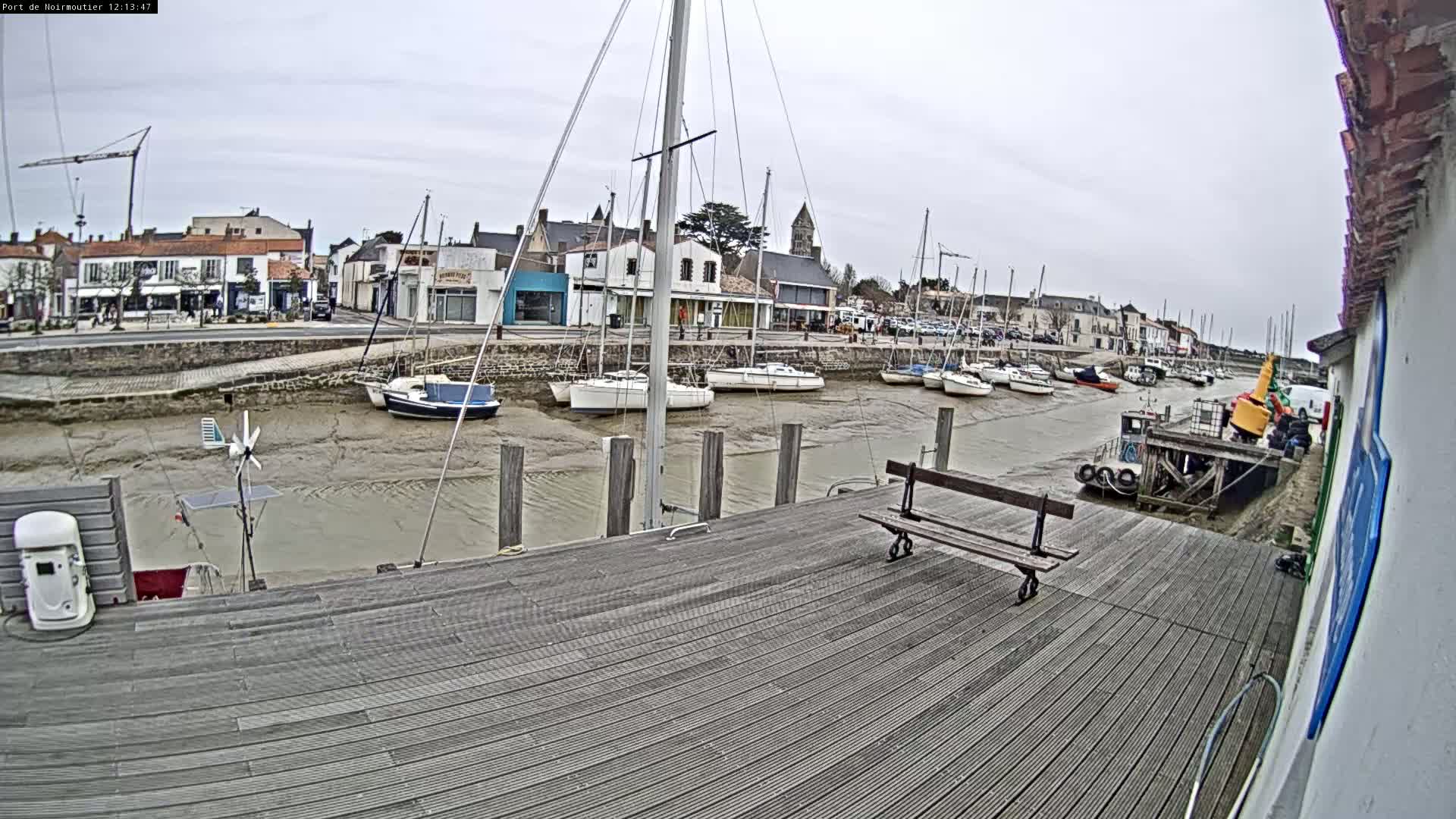 A wooden dock overlooks a harbor at low tide, where several sailboats are moored near a small town under an overcast sky.