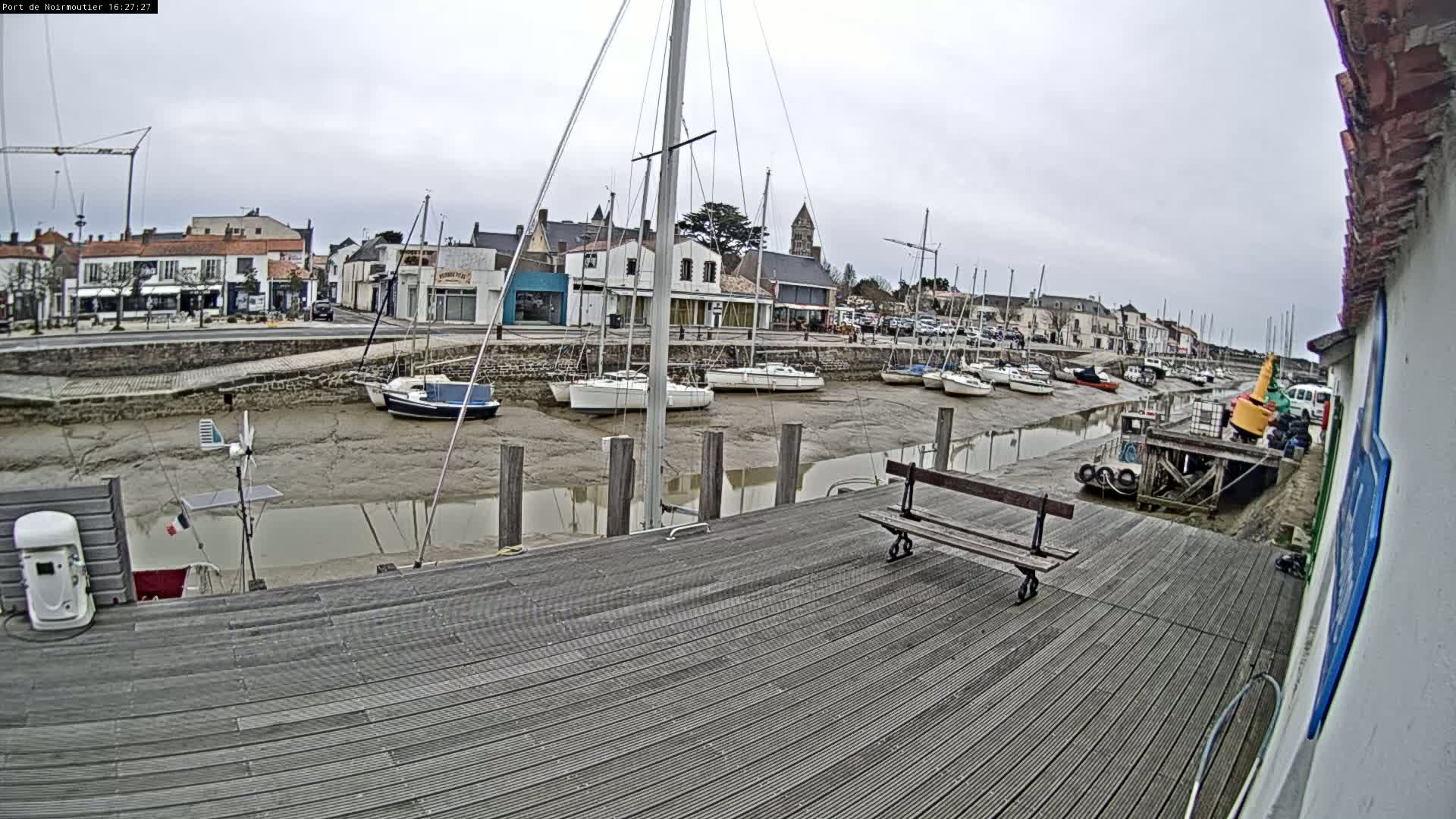 A wooden dock overlooks a calm harbor with several sailboats at low tide under an overcast sky.
