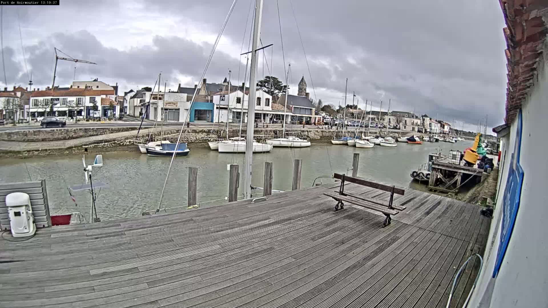 A wooden dock extends over calm water, lined with sailboats and a small town visible in the background under a cloudy sky.