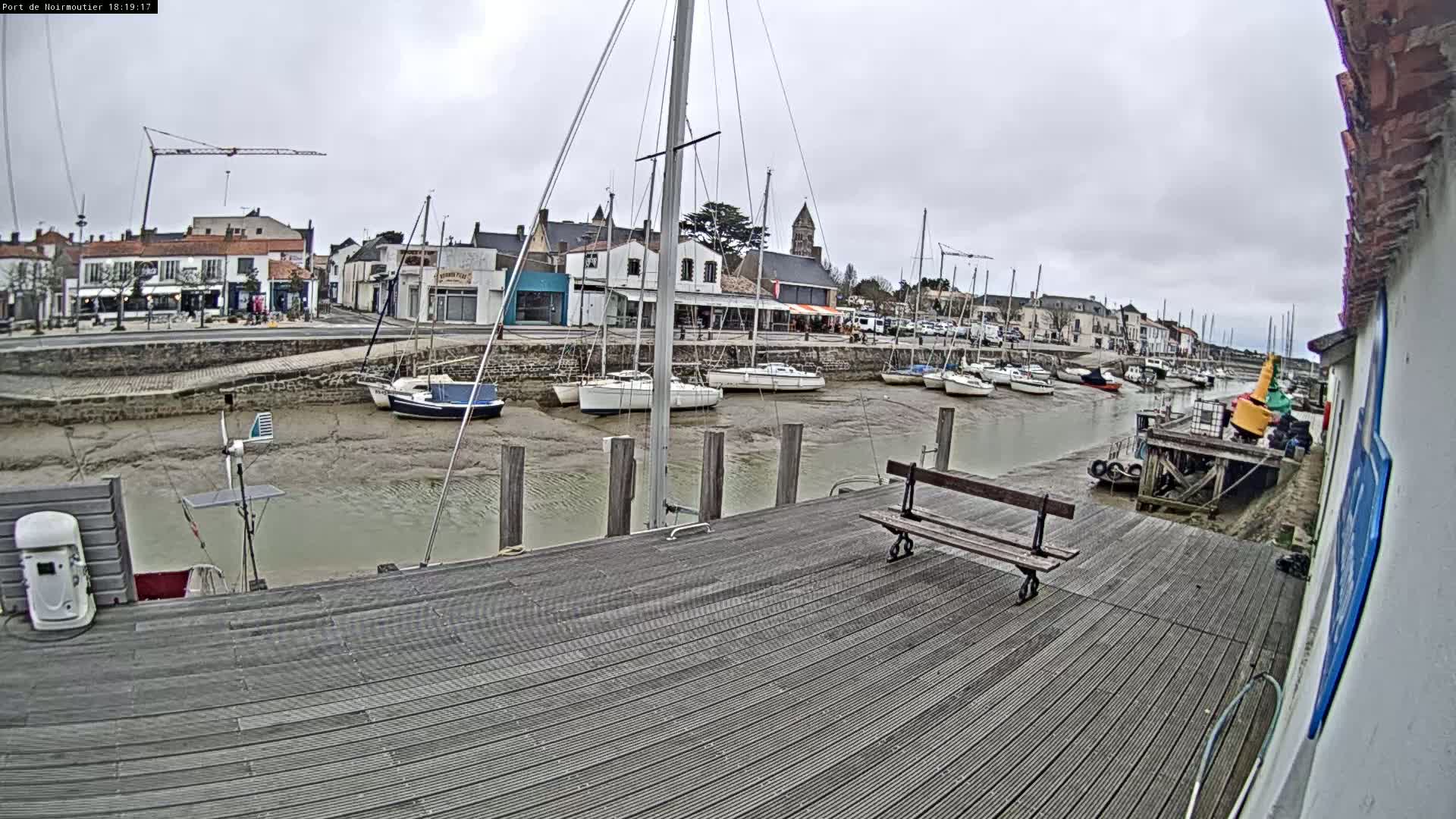 A wooden dock overlooks a harbor at low tide, where several sailboats are moored near a small town under a cloudy sky.
