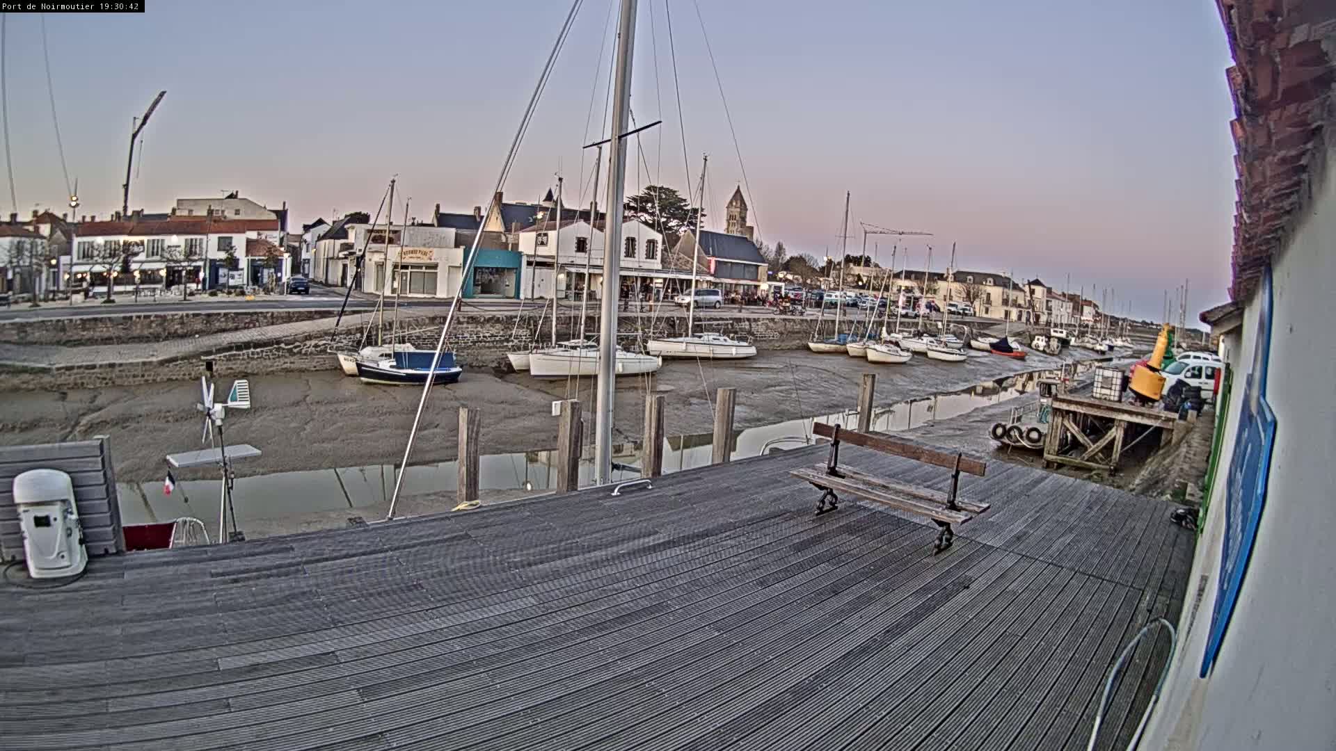A wooden dock overlooks a harbor at low tide, where several sailboats are moored near a small town under a twilight sky.