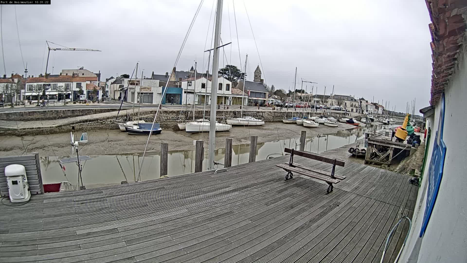 A wooden dock overlooks a calm harbor with numerous sailboats at low tide under an overcast sky.