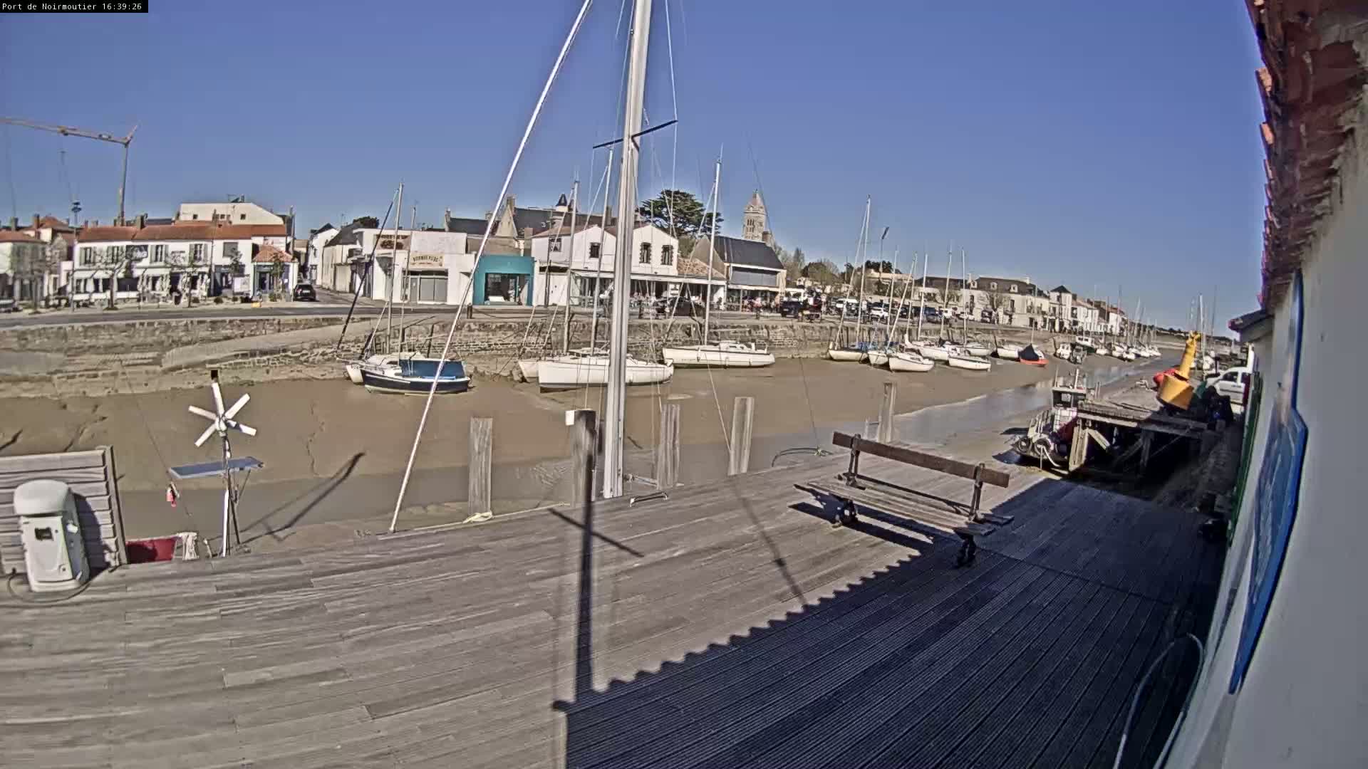 A sunny day at a harbor shows sailboats docked in shallow, muddy water near a town with buildings along the waterfront.