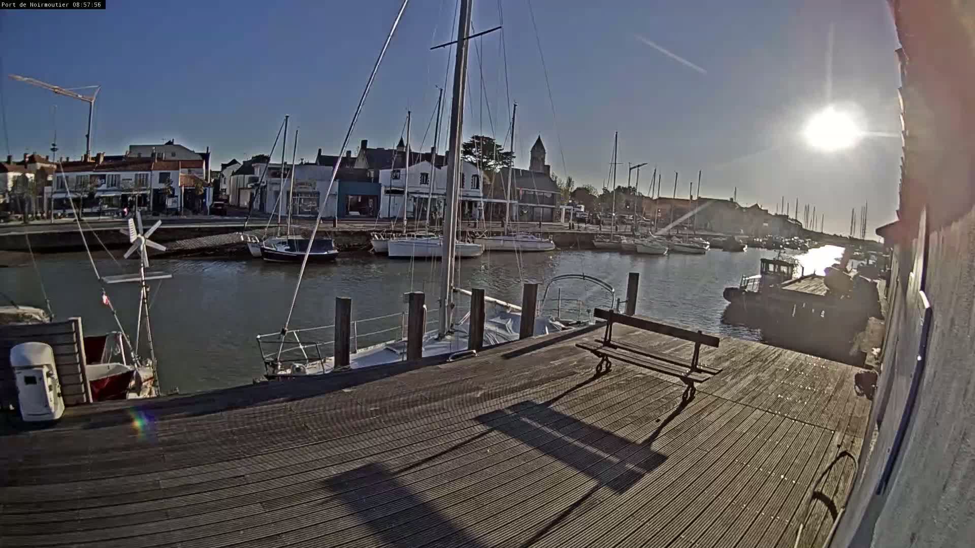 A sunny day at a harbor shows sailboats docked along a waterfront with buildings and a wooden dock in the foreground, featuring a park bench.