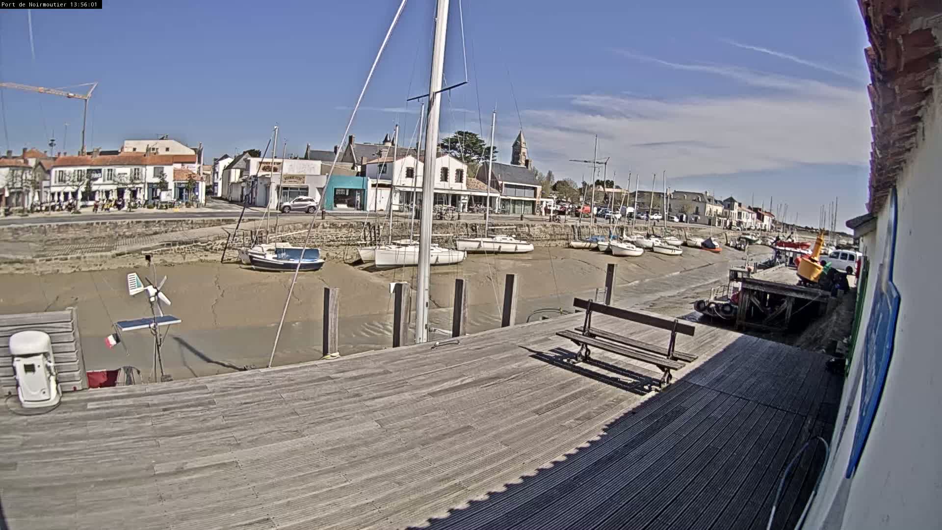 A sunny day at a harbor shows several sailboats docked in shallow, muddy water, with a wooden dock and a town in the background.